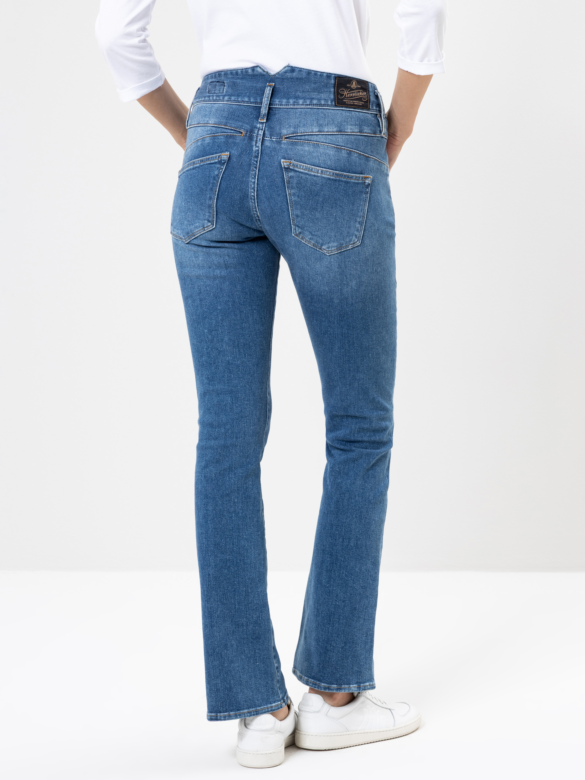 Mit dem Rücken zur Kamera stehende Person in hellblauen Jeans, weißem langärmeligen Hemd und weißen Turnschuhen in einer gut beleuchteten, minimalistischen Studioumgebung.