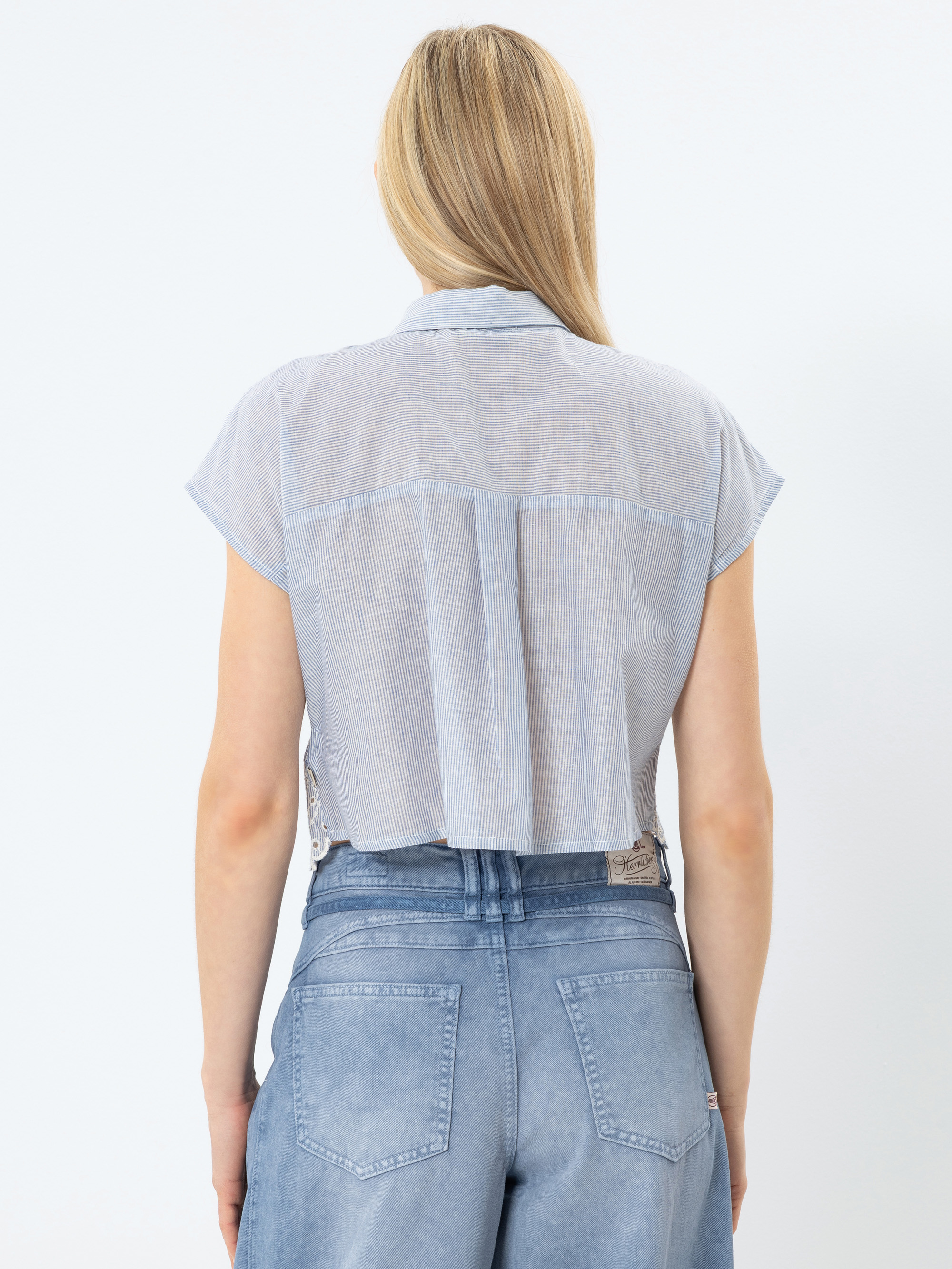 Eine Frau mit langen blonden Haaren, die ein kurzärmeliges, hellblaues Button-up-Hemd und eine hellblaue Jeans mit hoher Taille trägt, wird von hinten vor einem schlichten weißen Hintergrund gezeigt.