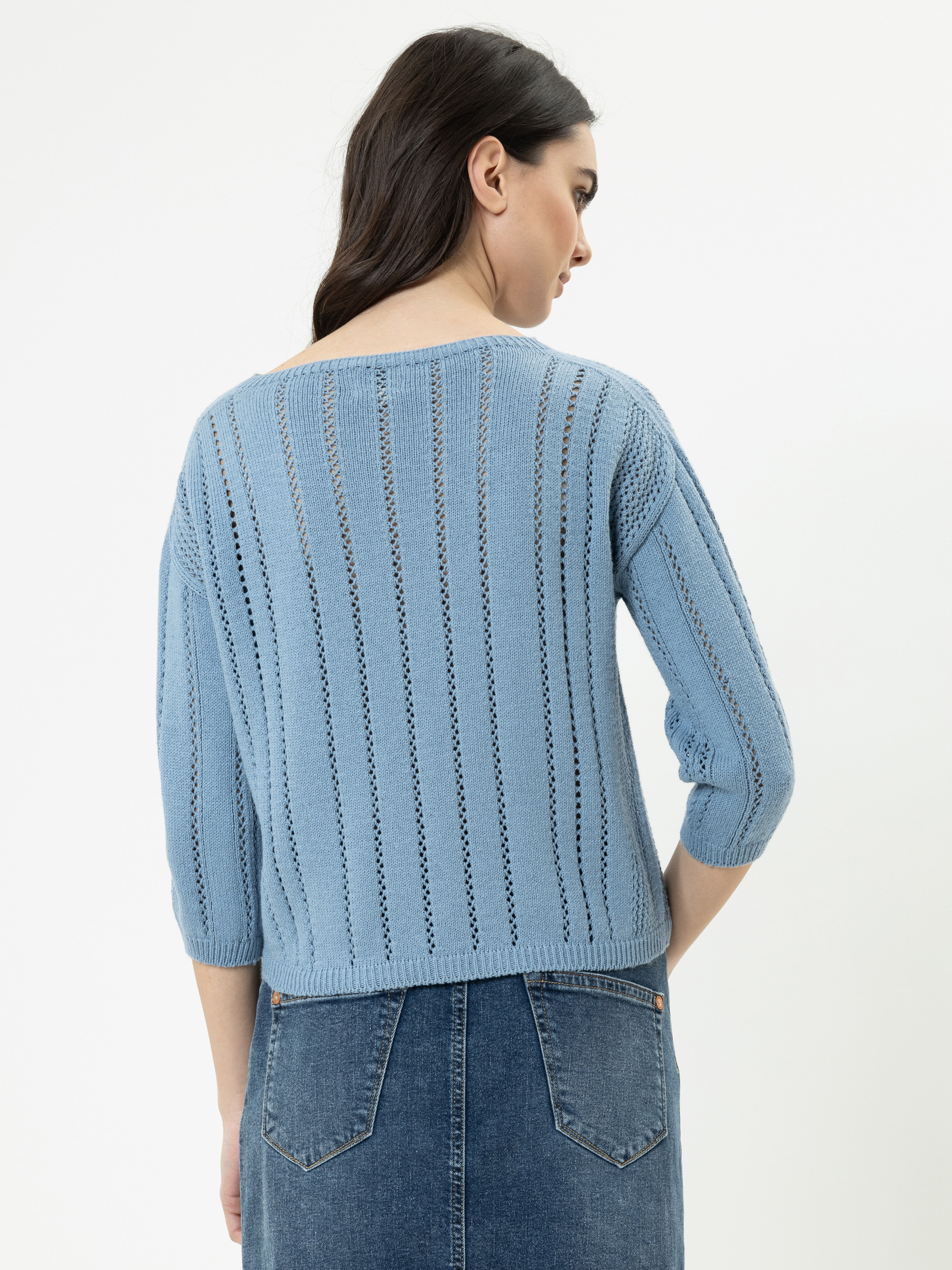 Frau mit langen dunklen Haaren, die einen hellblauen Strickpullover mit vertikalen Ösenstreifen und einen blauen Jeansrock trägt, steht mit dem Rücken zur Kamera vor einem schlichten weißen Hintergrund.