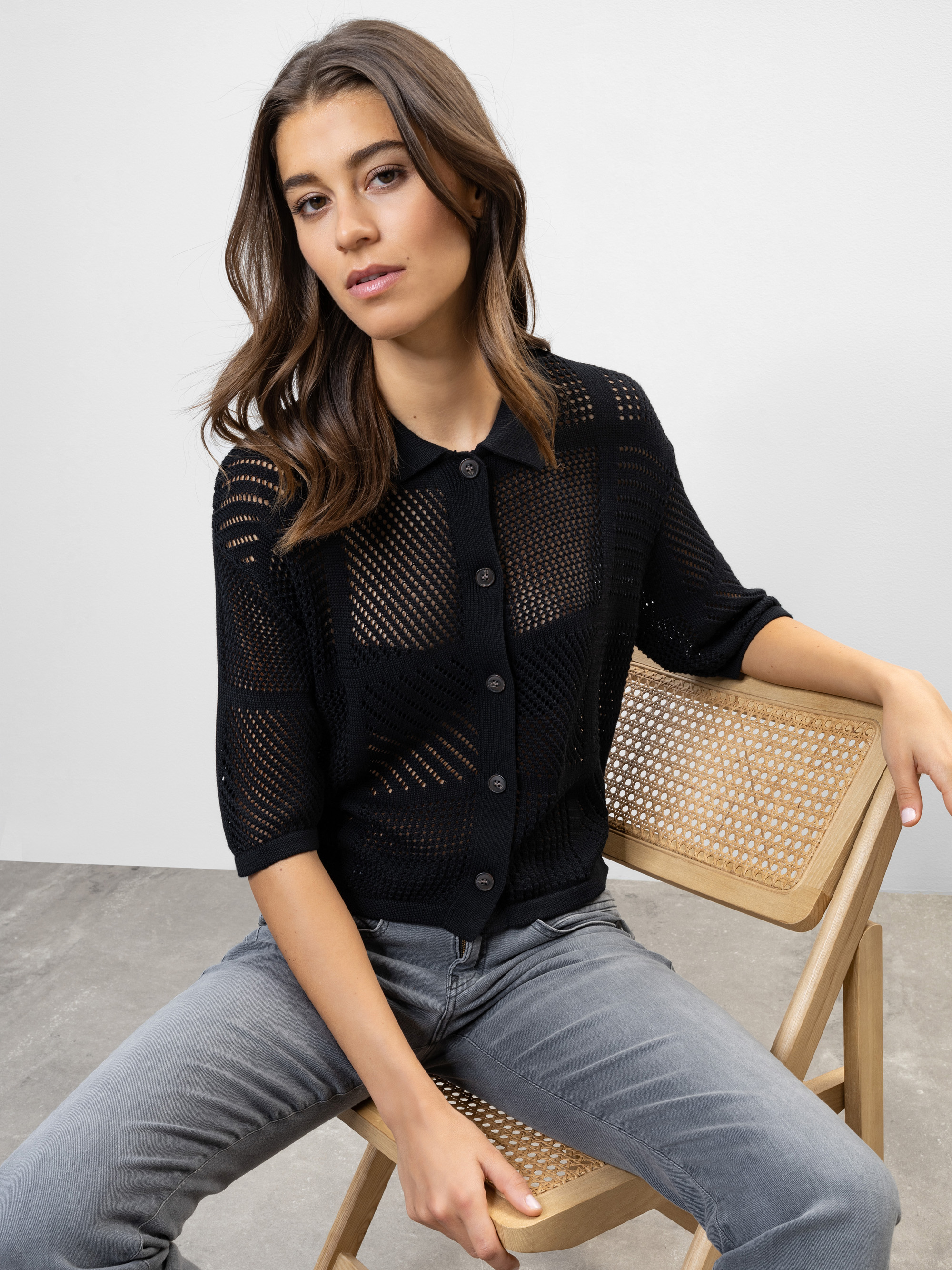 Eine Frau mit langen braunen Haaren sitzt seitlich auf einem Holzstuhl und trägt ein schwarzes, kurzärmeliges, geknöpftes Strickoberteil und graue Jeans vor einem schlichten hellgrauen Hintergrund.