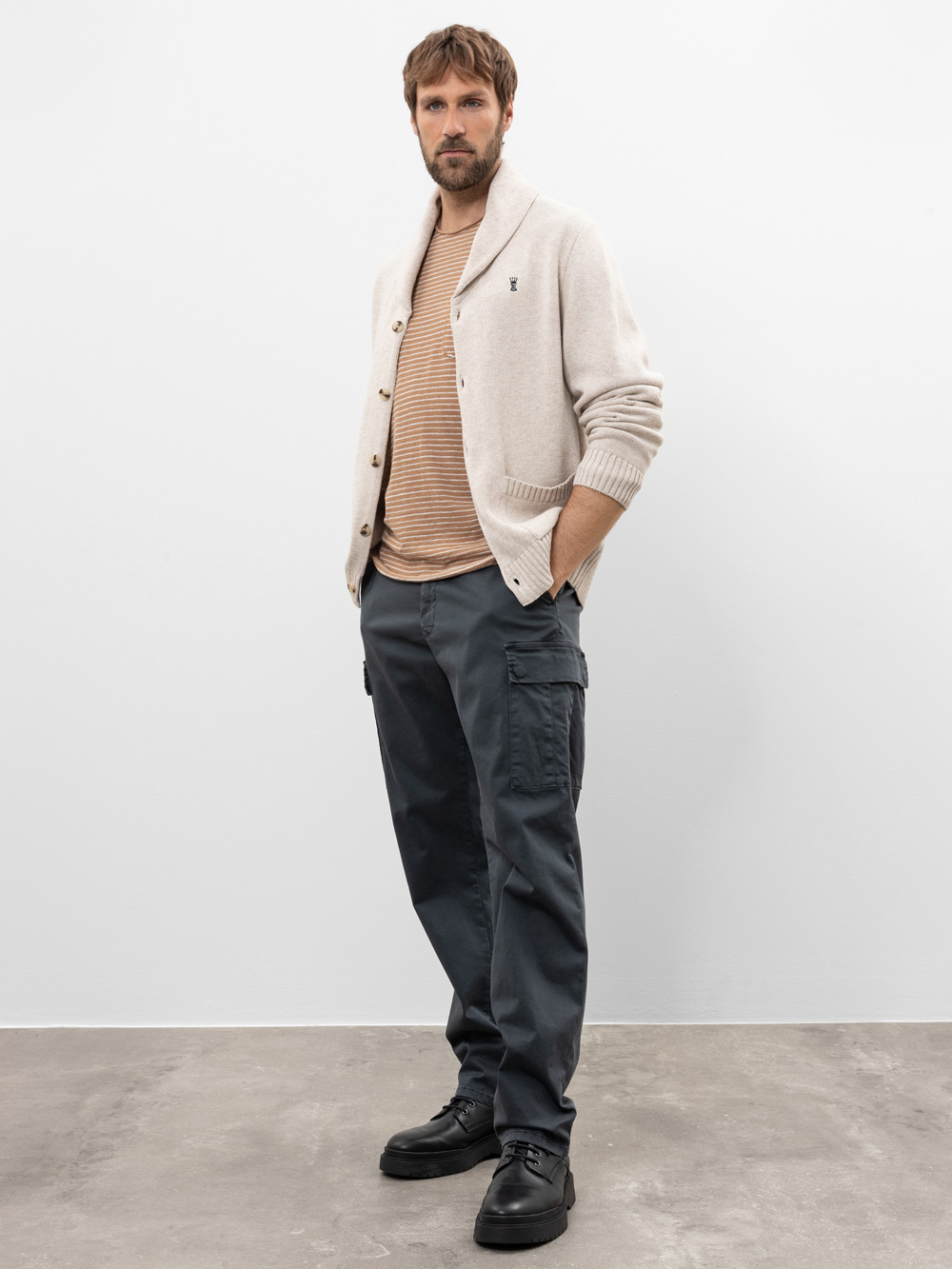 Ein Mann steht vor einer schlichten weißen Wand und trägt eine beigefarbene Strickjacke über einem gestreiften hellbraunen Hemd, eine dunkelgraue Cargohose und schwarze Schuhe. Er hat kurzes braunes Haar, einen Bart und die Hände in den Taschen.