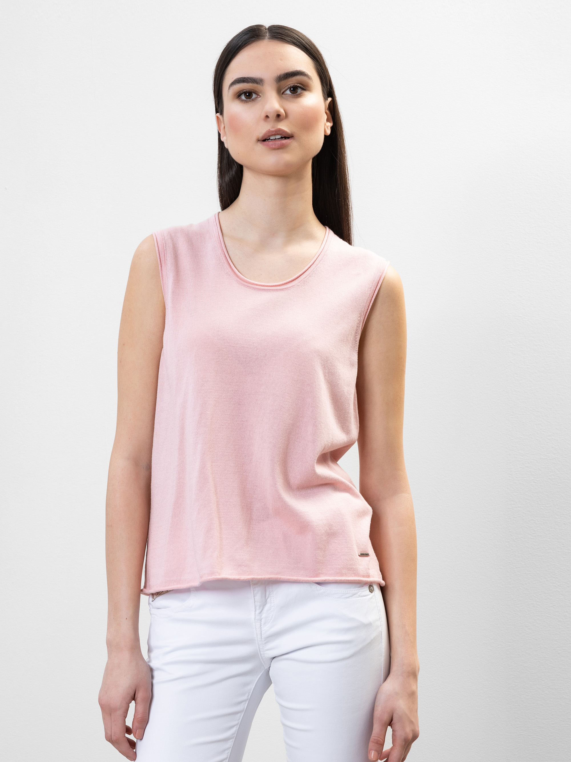Eine Frau mit langem glattem Haar, die ein ärmelloses hellrosa Oberteil und eine weiße Hose trägt, steht vor einem schlichten hellgrauen Hintergrund und schaut mit neutralem Blick leicht zur Seite.