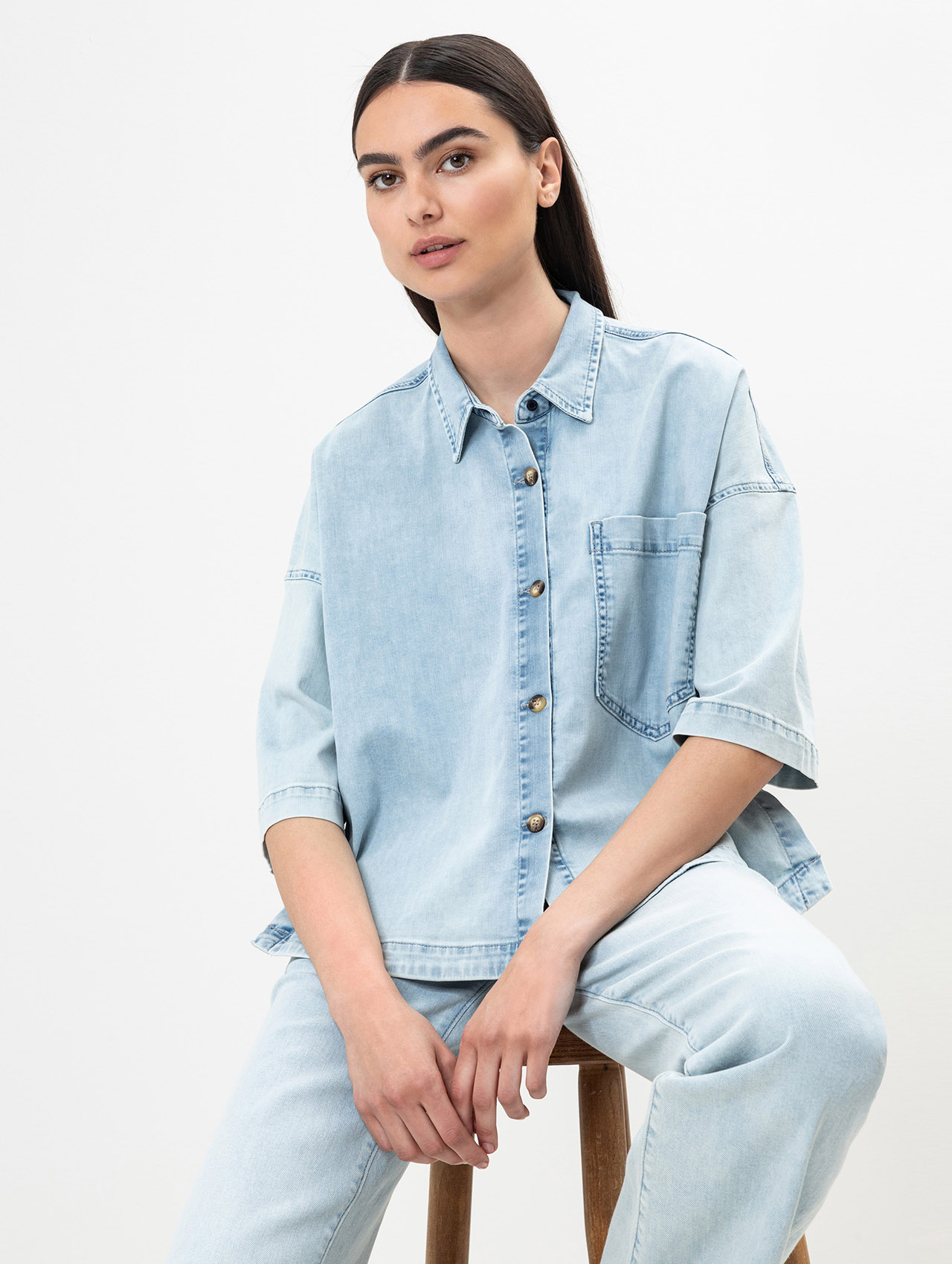 Eine Frau mit langen dunklen Haaren sitzt auf einem Holzschemel, trägt ein hellblaues Jeanshemd mit hochgekrempelten Ärmeln und eine dazu passende hellblaue Jeans, posiert vor einem schlichten weißen Hintergrund.