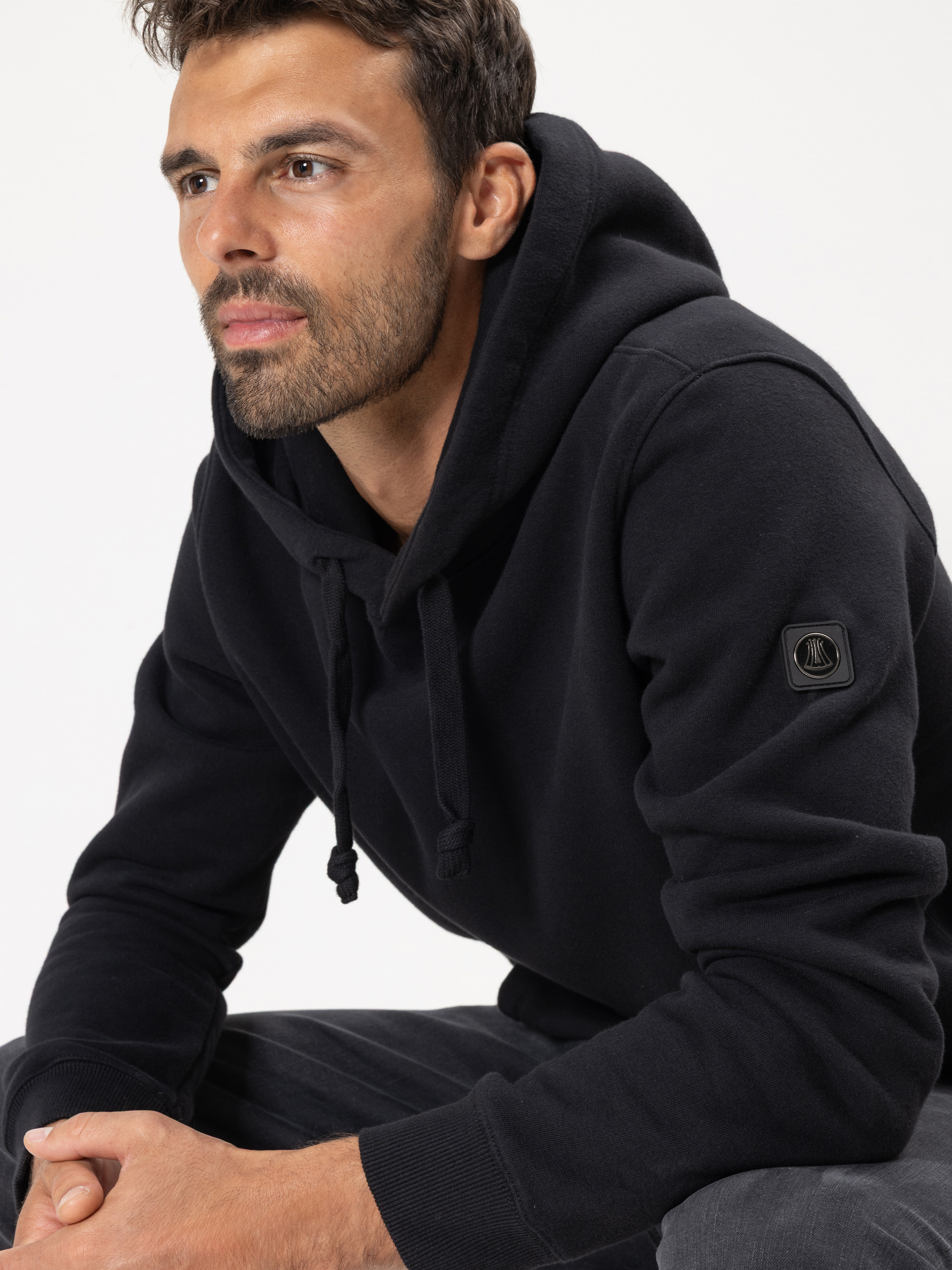 Ein Mann mit kurzen dunklen Haaren und einem Bart trägt ein schwarzes Kapuzensweatshirt mit einem kleinen Aufnäher am Ärmel. Er sitzt und schaut nachdenklich in die Ferne vor einem schlichten Hintergrund.