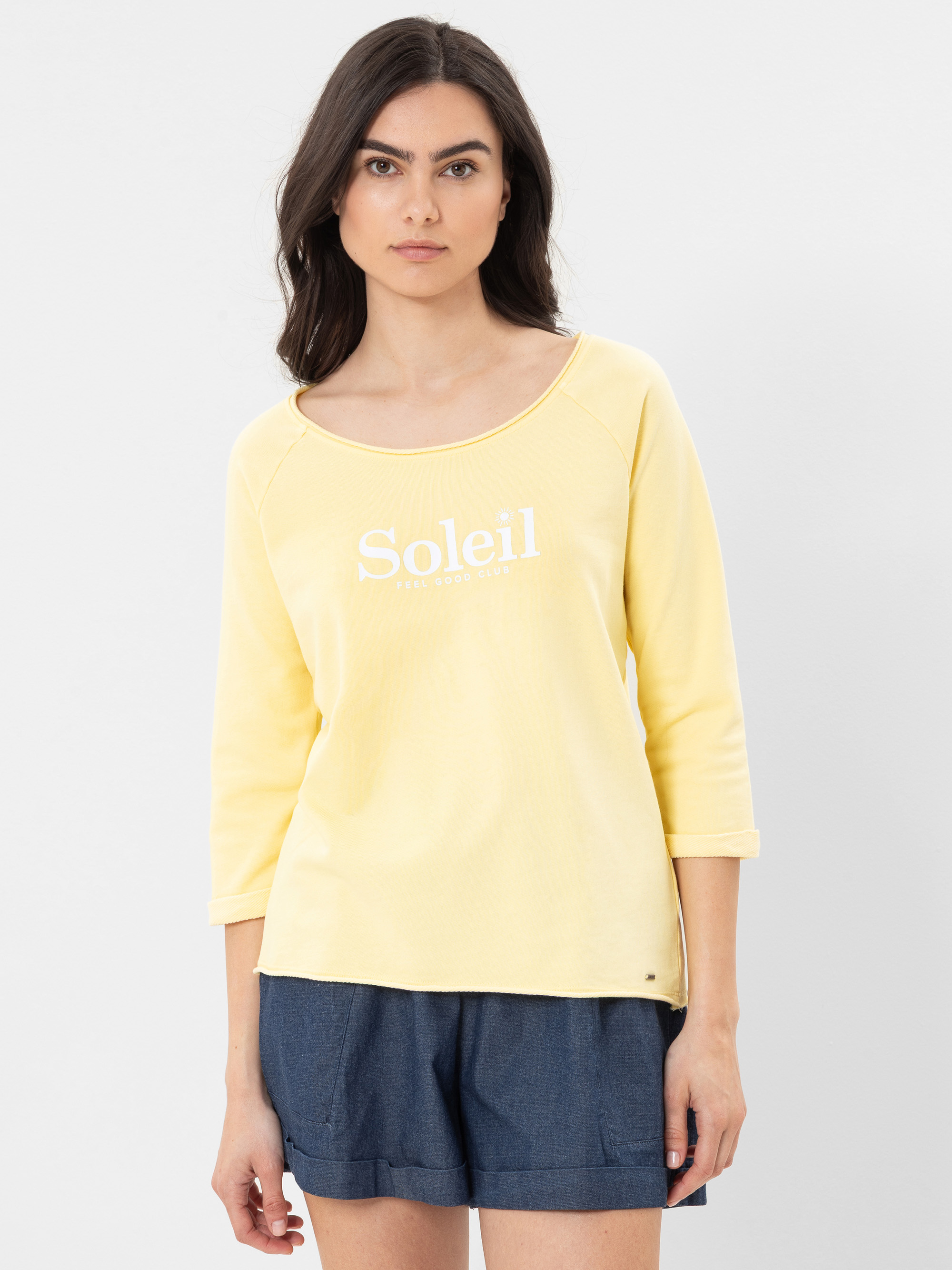 Eine Frau mit langen dunklen Haaren trägt ein hellgelbes Hemd mit der Aufschrift "Soleil" in weißen Buchstaben und dunkelblaue Shorts. Sie steht vor einem schlichten weißen Hintergrund und blickt mit neutraler Miene nach vorne.