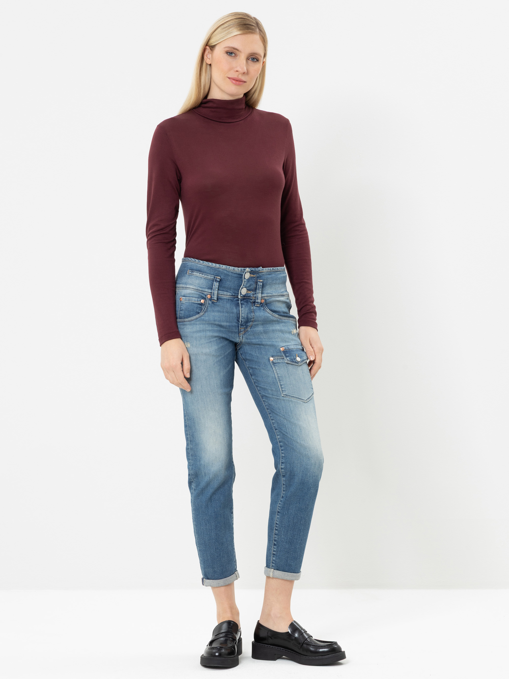 Eine Person steht vor einem weißen Hintergrund und trägt einen burgunderroten Rollkragenpullover, hellblaue Jeans mit Umschlag und Taschen sowie schwarze Slipper. Sie hat langes blondes Haar und einen entspannten Gesichtsausdruck.