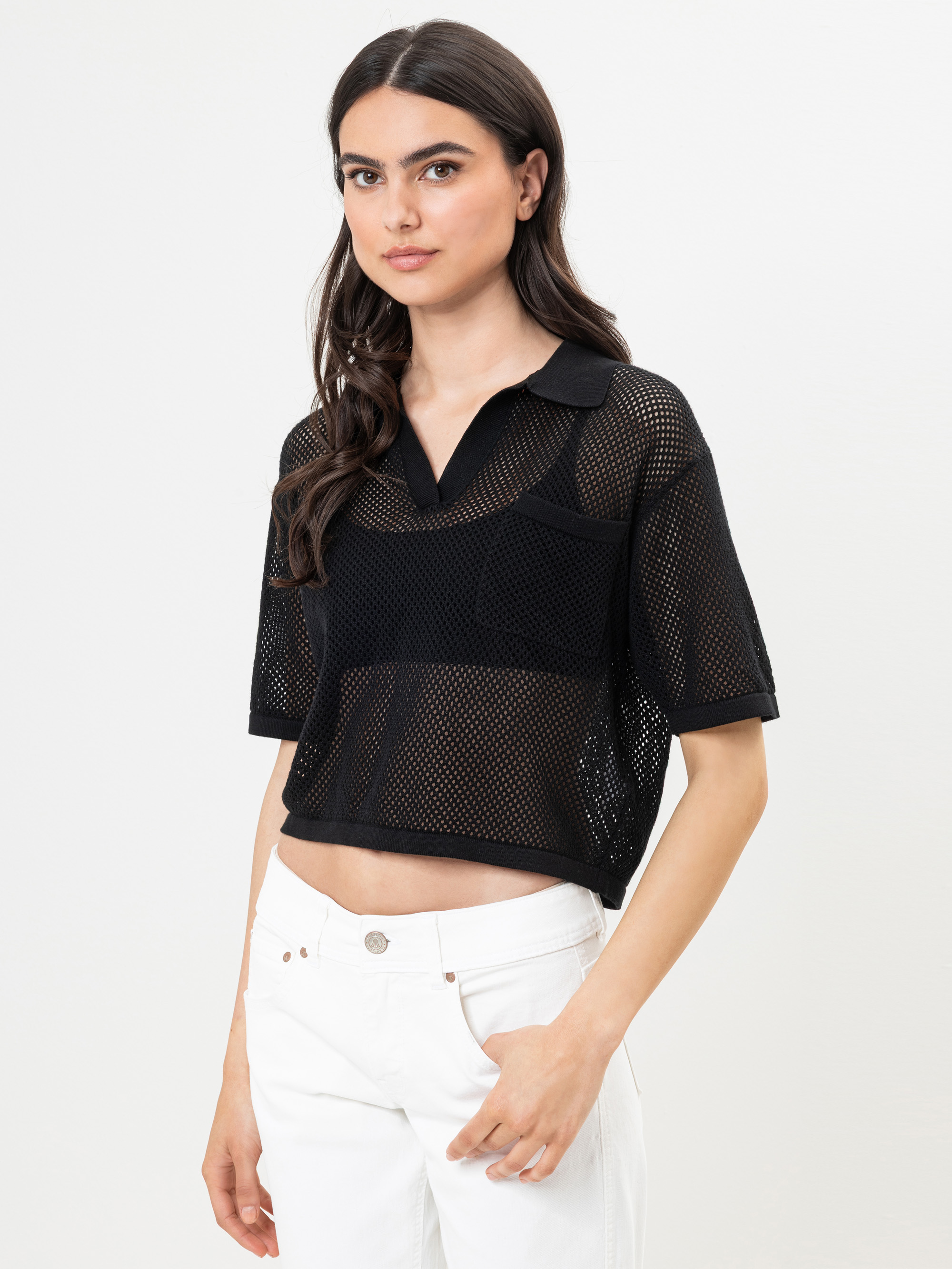 Eine Frau mit langen dunklen Haaren trägt ein schwarzes, kurzärmeliges Netz-Poloshirt über einem schwarzen Oberteil und einer weißen Hose und steht vor einem schlichten weißen Hintergrund.