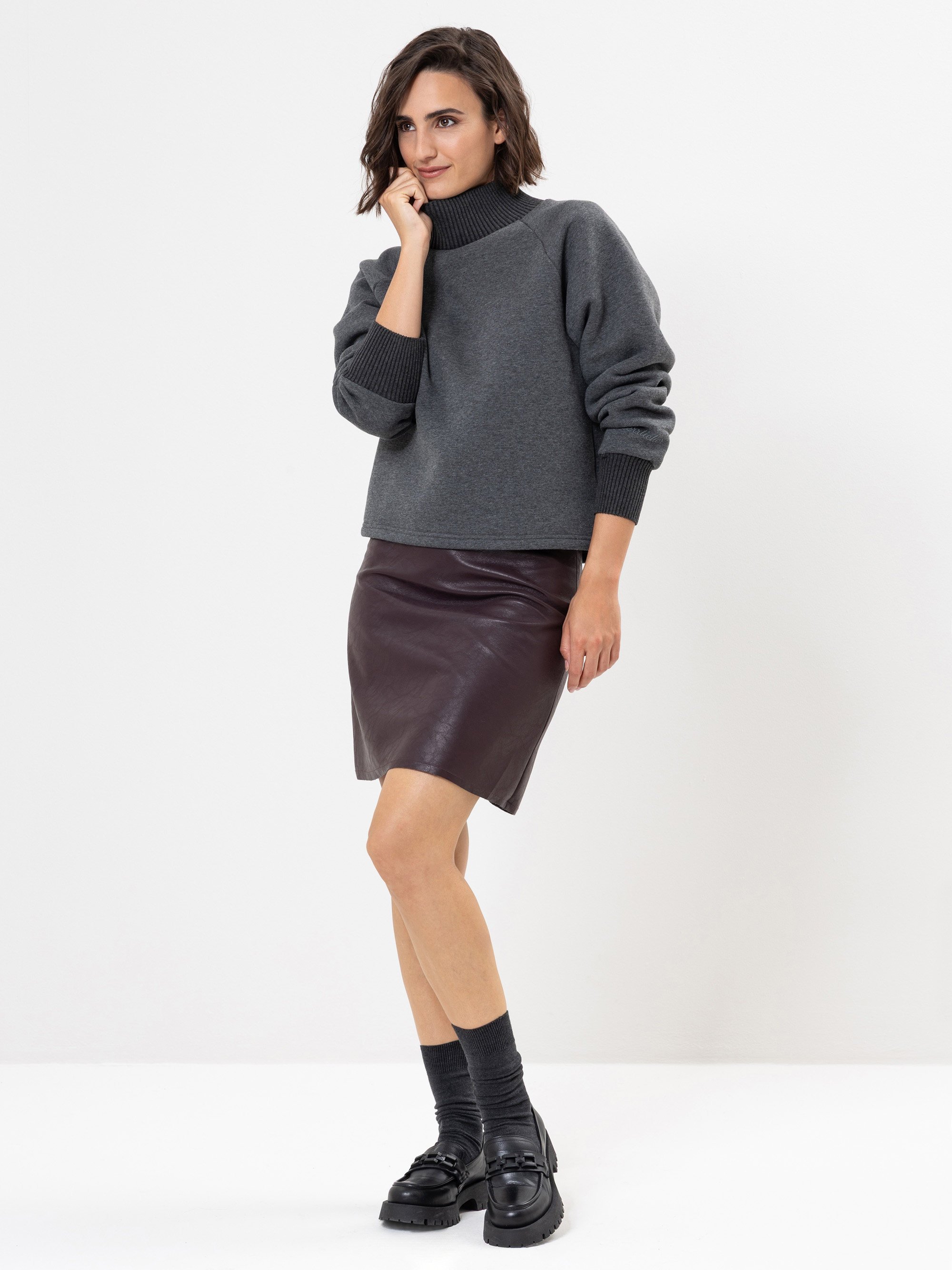 Frau, die vor einem weißen Hintergrund steht und einen dunkelgrauen Rollkragenpullover, einen dunkelbraunen Lederrock, graue Socken und schwarze klobige Schuhe trägt, wobei eine Hand ihr Kinn berührt und sie leicht lächelt.