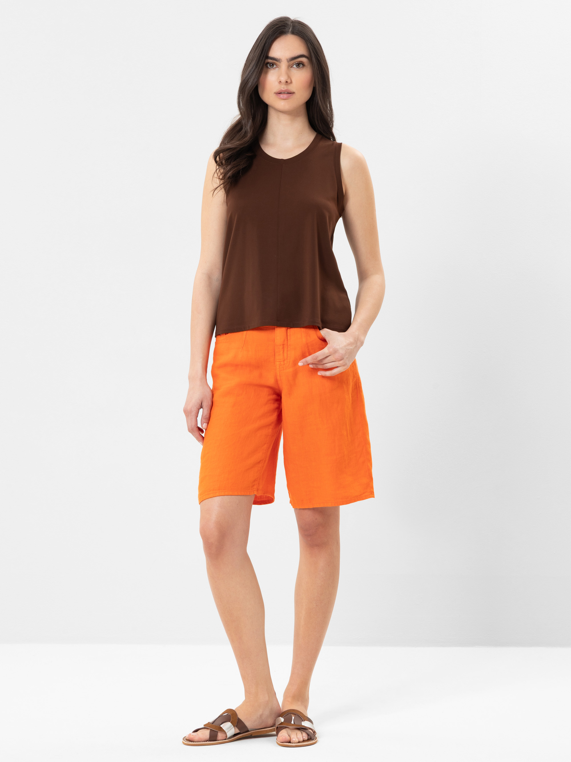 Eine Frau steht vor einem schlichten Hintergrund und trägt ein ärmelloses braunes Oberteil, leuchtend orangefarbene knielange Shorts und braune Sandalen. Sie hat langes dunkles Haar und eine Hand in der Tasche.