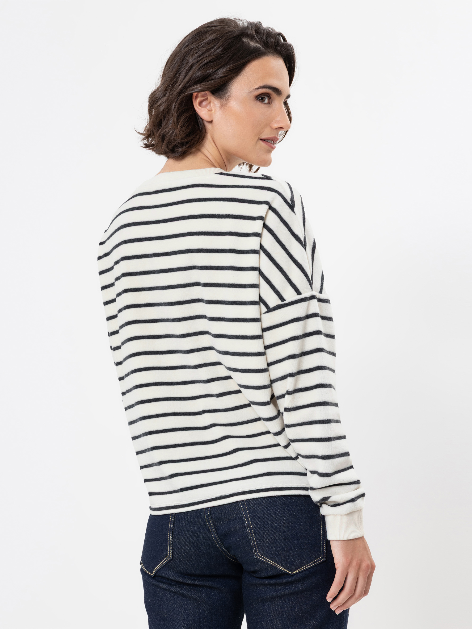 Eine Frau mit kurzen braunen Haaren trägt einen weißen Pullover mit schwarzen Querstreifen und dunkelblaue Jeans. Sie steht und schaut über ihre Schulter vor einem schlichten weißen Hintergrund.