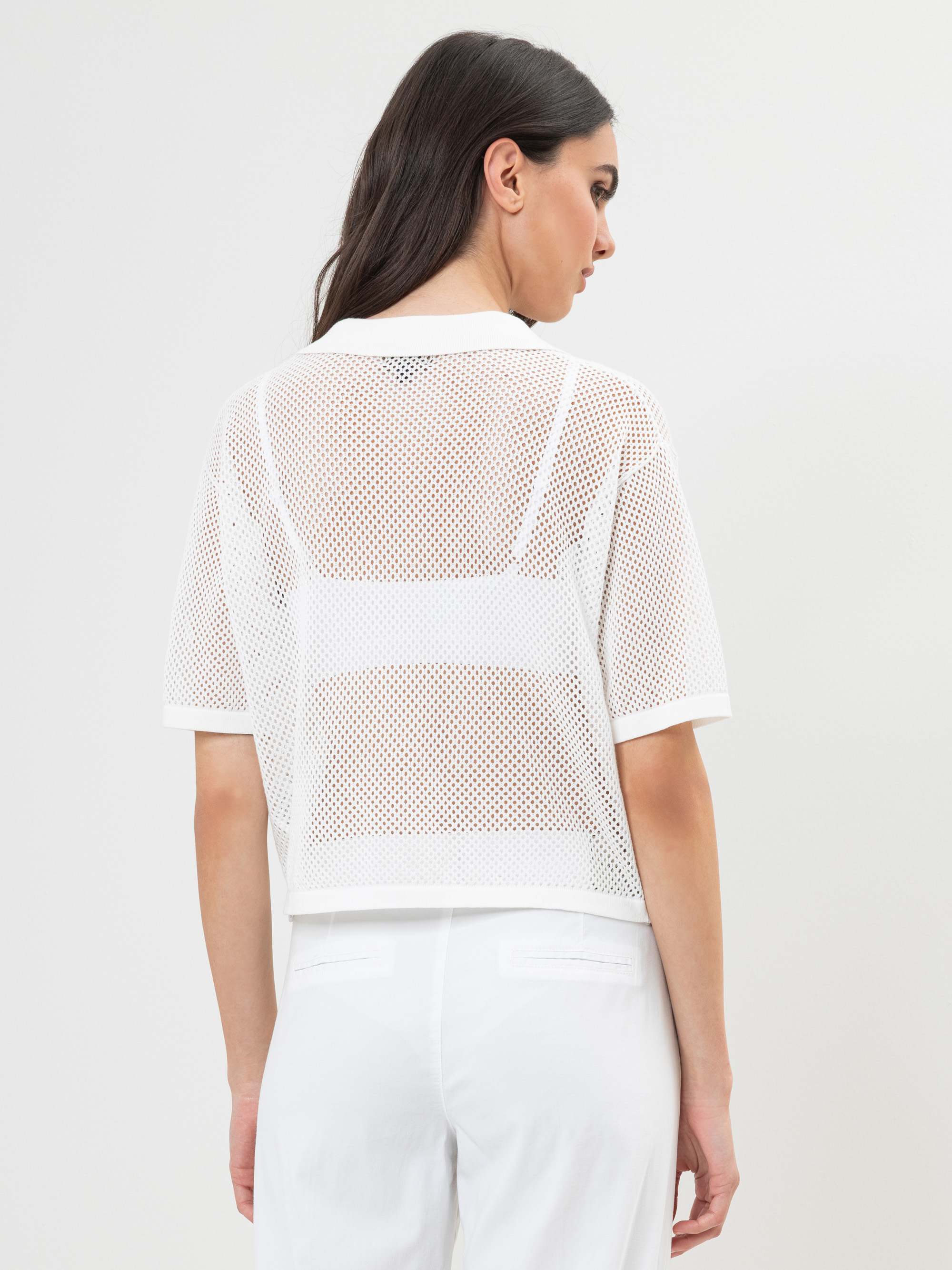 Eine Frau mit langen dunklen Haaren wird von hinten gezeigt. Sie trägt ein weißes kurzärmeliges Netzoberteil über einem weißen Bralette und eine weiße Hose vor einem schlichten hellen Hintergrund.