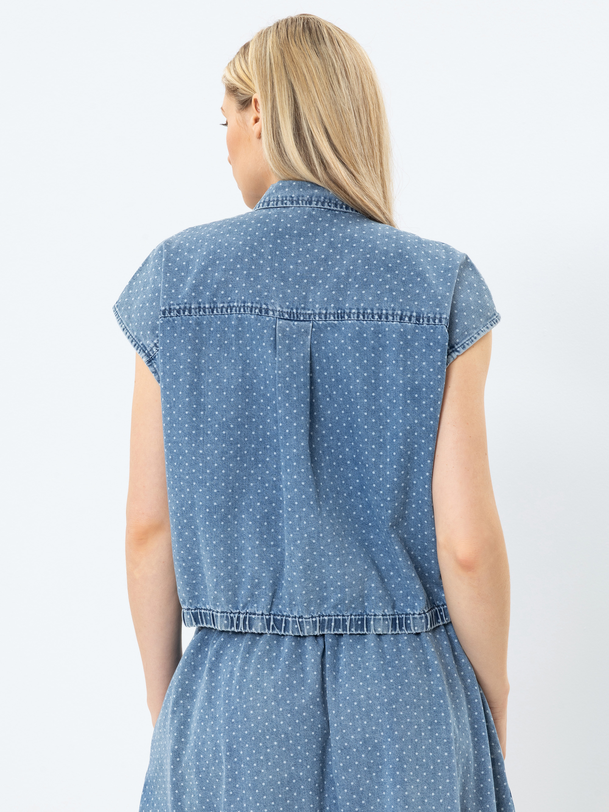 Eine Frau mit langen blonden Haaren ist von hinten zu sehen. Sie trägt ein ärmelloses blaues Jeanskleid mit kleinen weißen Punkten. Ihre Arme liegen entspannt an ihren Seiten vor einem schlichten weißen Hintergrund.