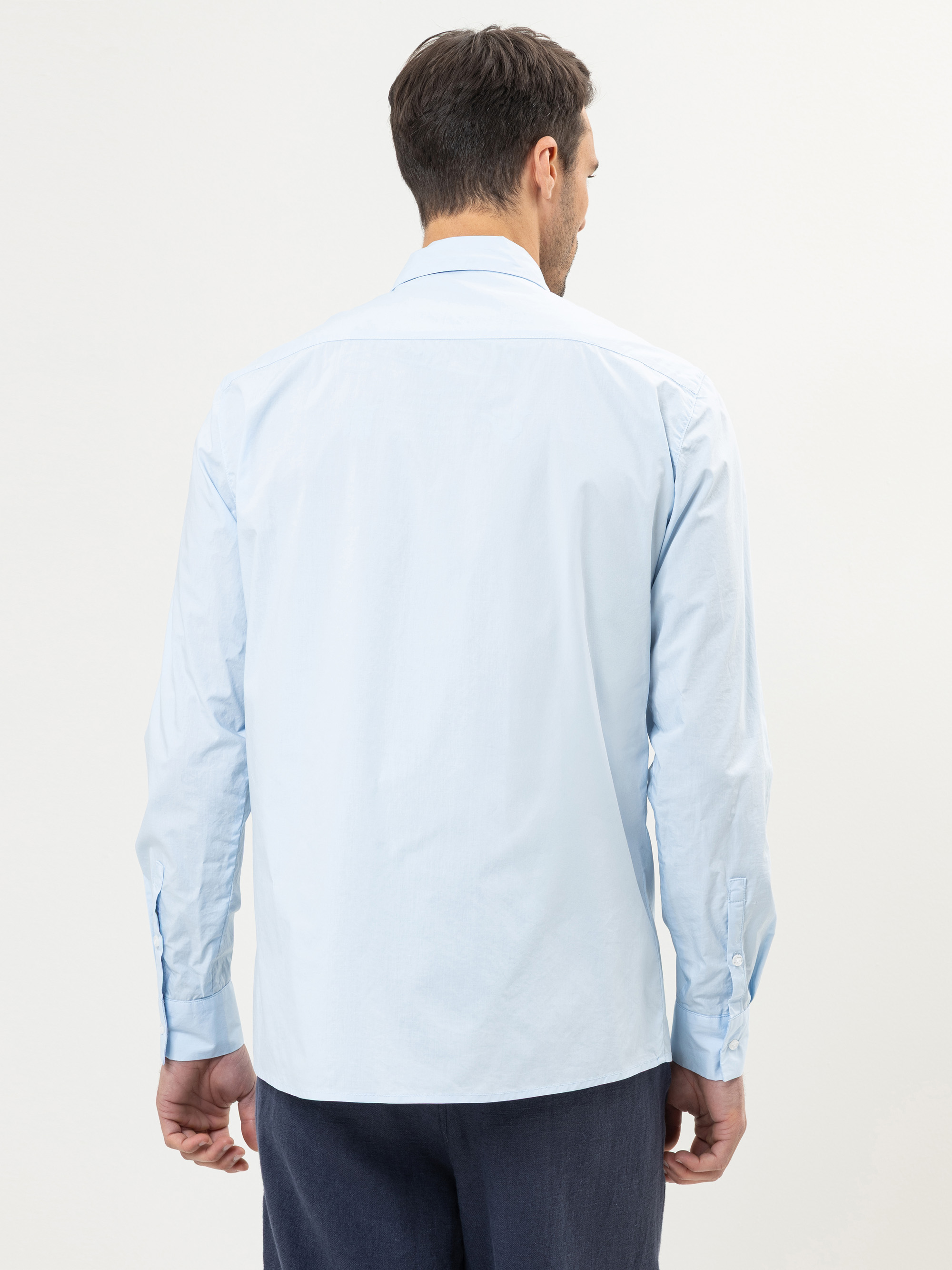 Ein Mann steht mit dem Rücken zur Kamera, trägt ein hellblaues, langärmeliges Hemd zum Aufknöpfen und eine dunkle Hose vor einem schlichten, hellen Hintergrund.