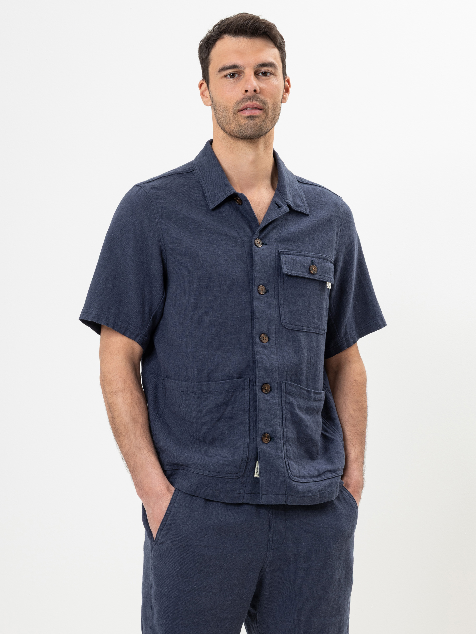 Ein Mann, der ein marineblaues, kurzärmeliges Hemd mit mehreren Taschen und eine dazu passende Hose trägt, steht mit den Händen in den Taschen vor einem schlichten weißen Hintergrund.