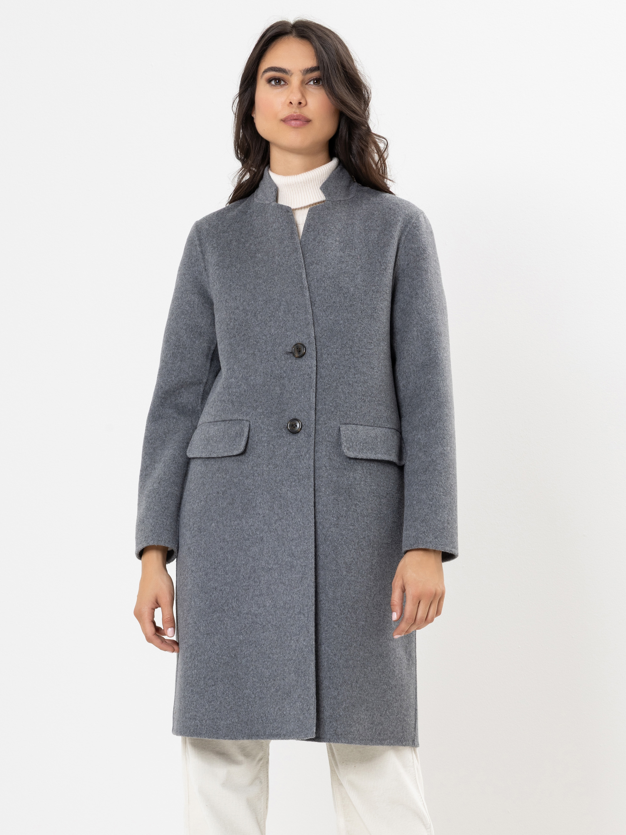 Eine Frau steht vor einem weißen Hintergrund und trägt einen grauen, knielangen Mantel mit zwei Knöpfen und Klappentaschen, einen cremefarbenen Rollkragenpullover und eine weiße Hose.