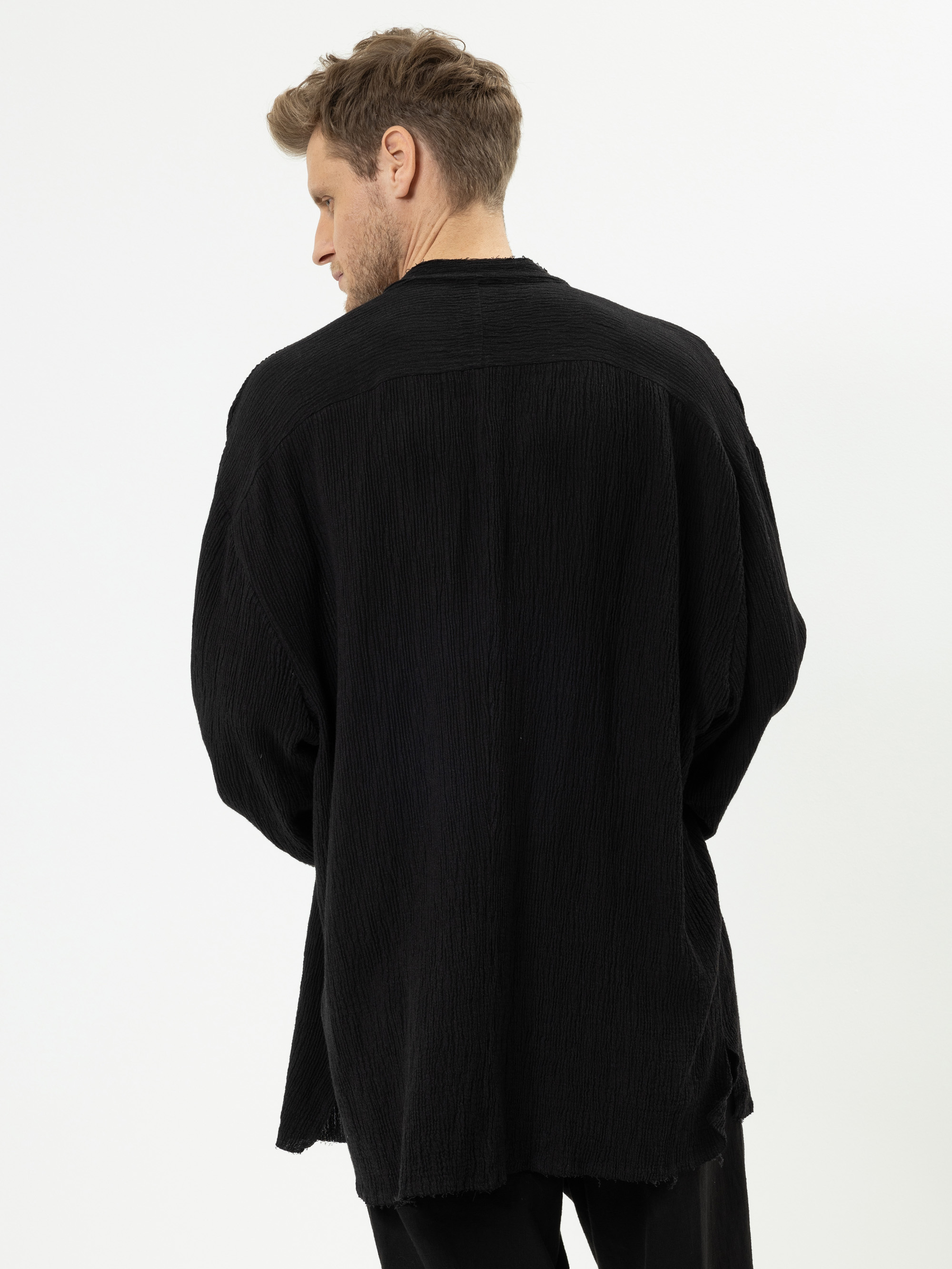 Ein Mann mit hellbraunem Haar steht mit dem Rücken zur Kamera und trägt ein locker sitzendes, langärmeliges schwarzes Hemd und eine schwarze Hose vor einem schlichten weißen Hintergrund.