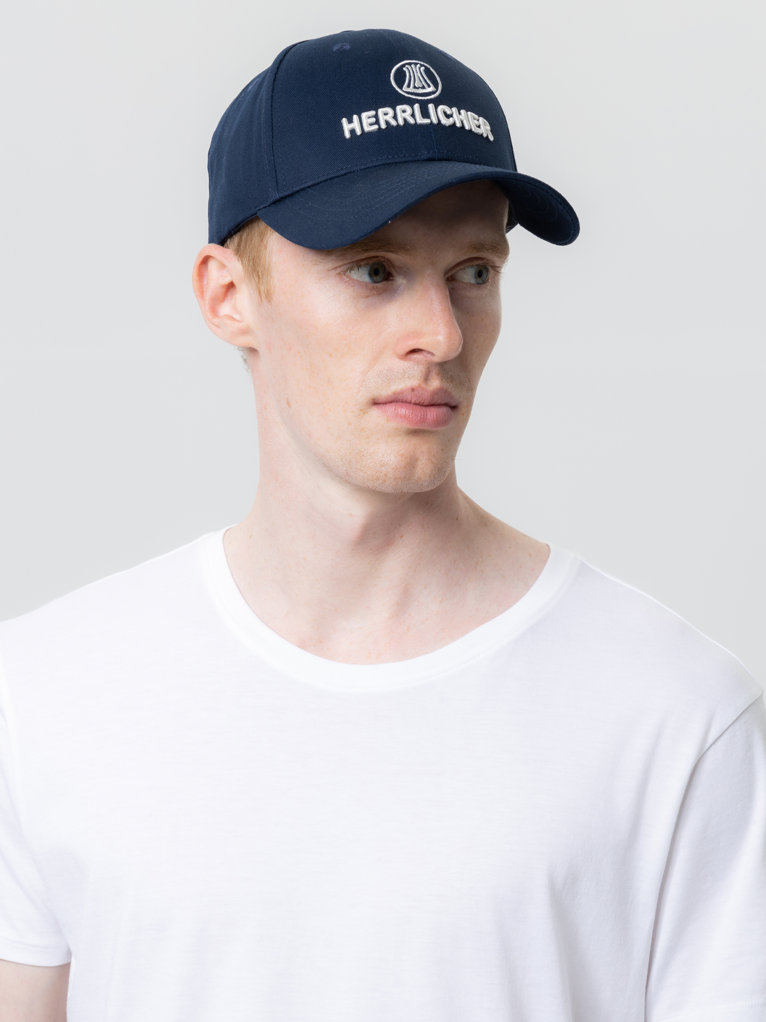 Eine Person, die eine marineblaue Herrlicher-Baseballmütze und ein schlichtes weißes T-Shirt trägt, blickt leicht nach links, vor einem hellgrauen Hintergrund.