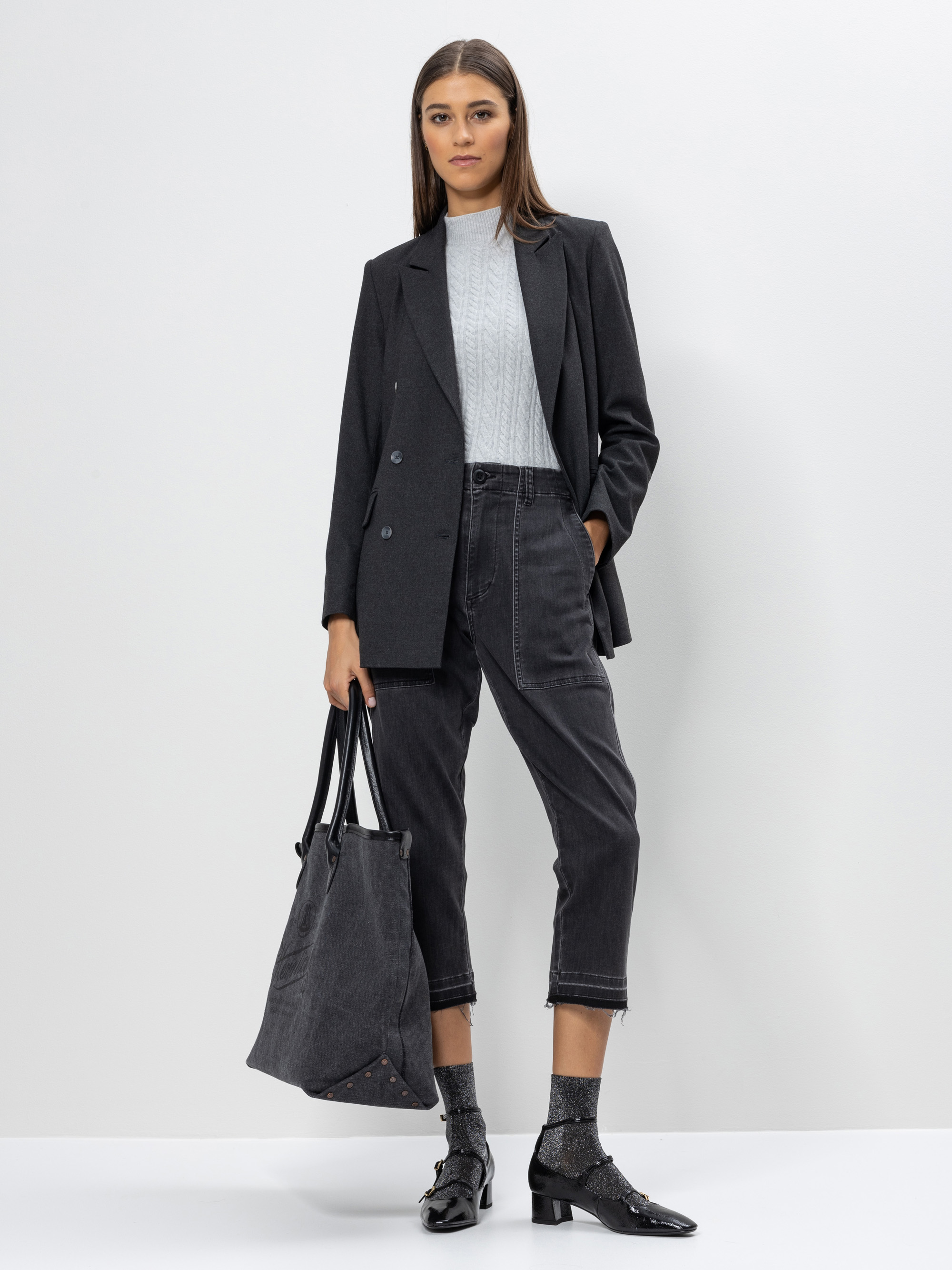 Eine Frau steht vor einem schlichten Hintergrund, trägt einen dunklen Blazer, einen hellgrauen Rollkragenpullover, eine schwarze Jeans, glitzernde Socken, schwarze Schuhe und hält eine große schwarze Tragetasche.