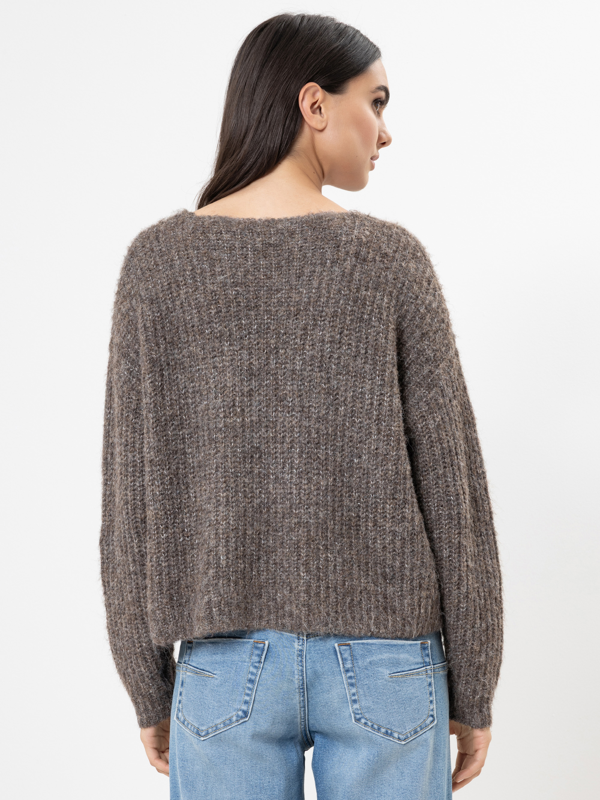 Eine Frau mit langen dunklen Haaren wird von hinten gezeigt. Sie trägt einen locker sitzenden, strukturierten braunen Pullover und hellblaue Jeans mit Gesäßtaschen und steht vor einem schlichten weißen Hintergrund.