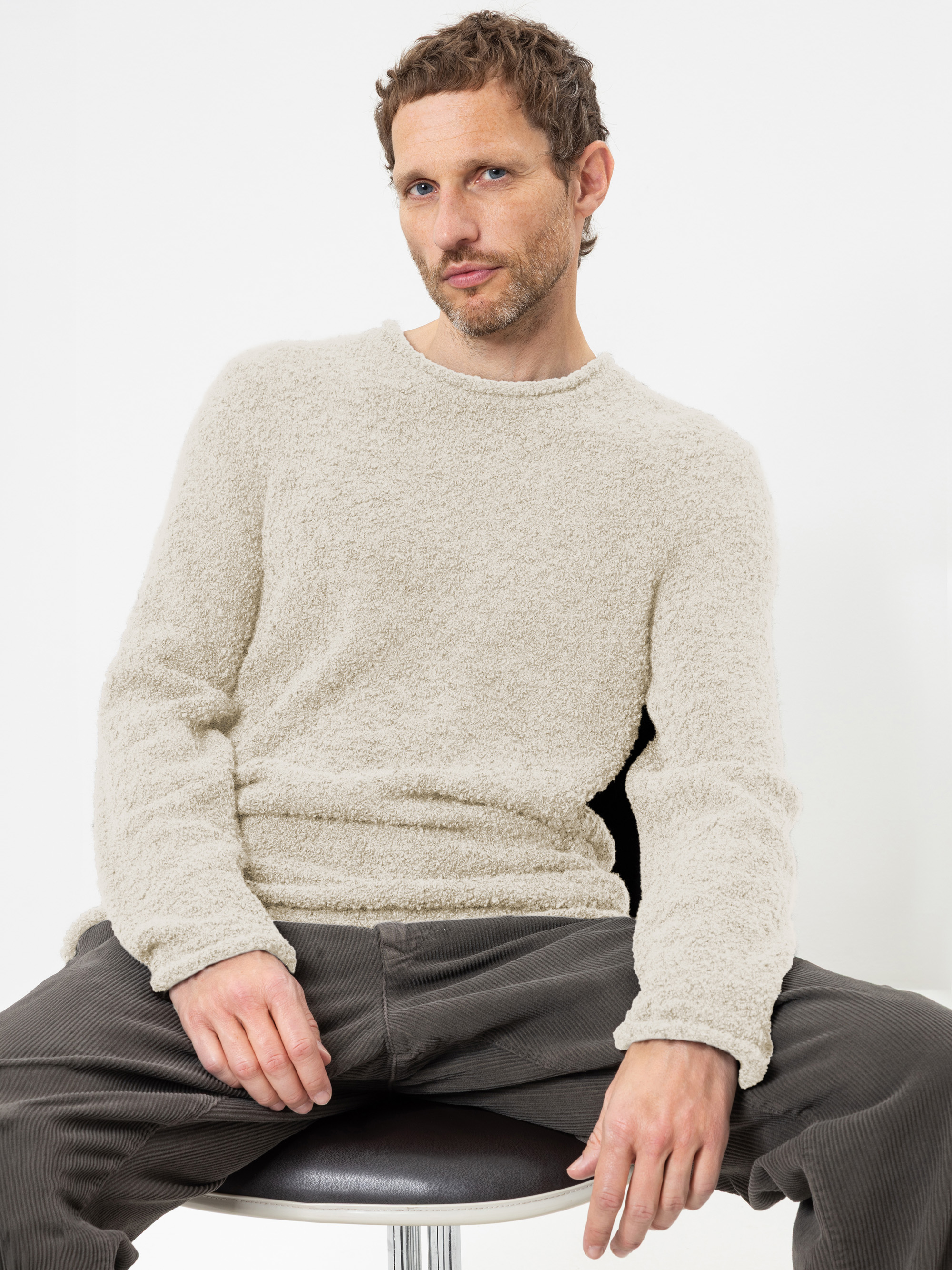 Ein Mann mit kurzen braunen Haaren und leichtem Bartwuchs sitzt auf einem Hocker, trägt einen strukturierten beigen Pullover und eine dunkle Hose und blickt mit ausdruckslosem Gesicht vor einem schlichten weißen Hintergrund in die Kamera.