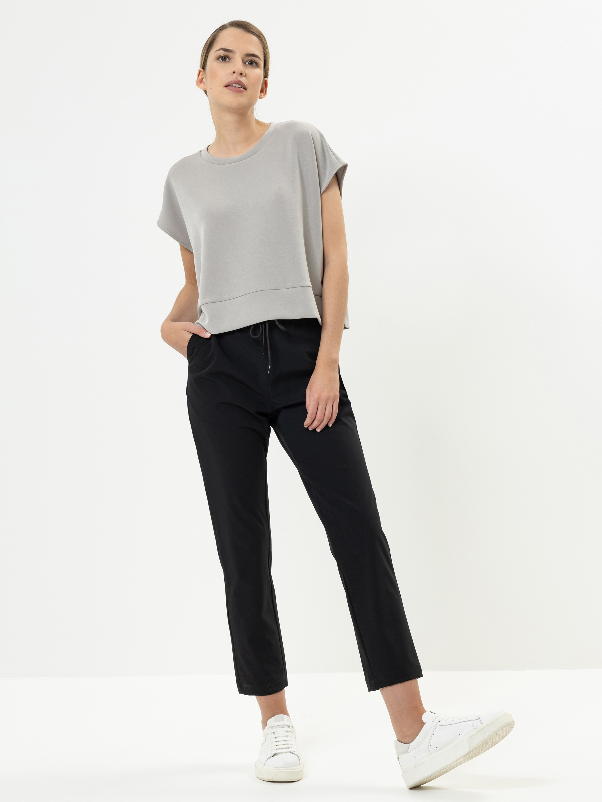Eine Frau steht vor einem weißen Hintergrund. Sie trägt ein hellgraues, kurzärmeliges, locker sitzendes Oberteil, eine schwarze Hose mit Kordelzug und weiße Turnschuhe und hat eine Hand in der Tasche.