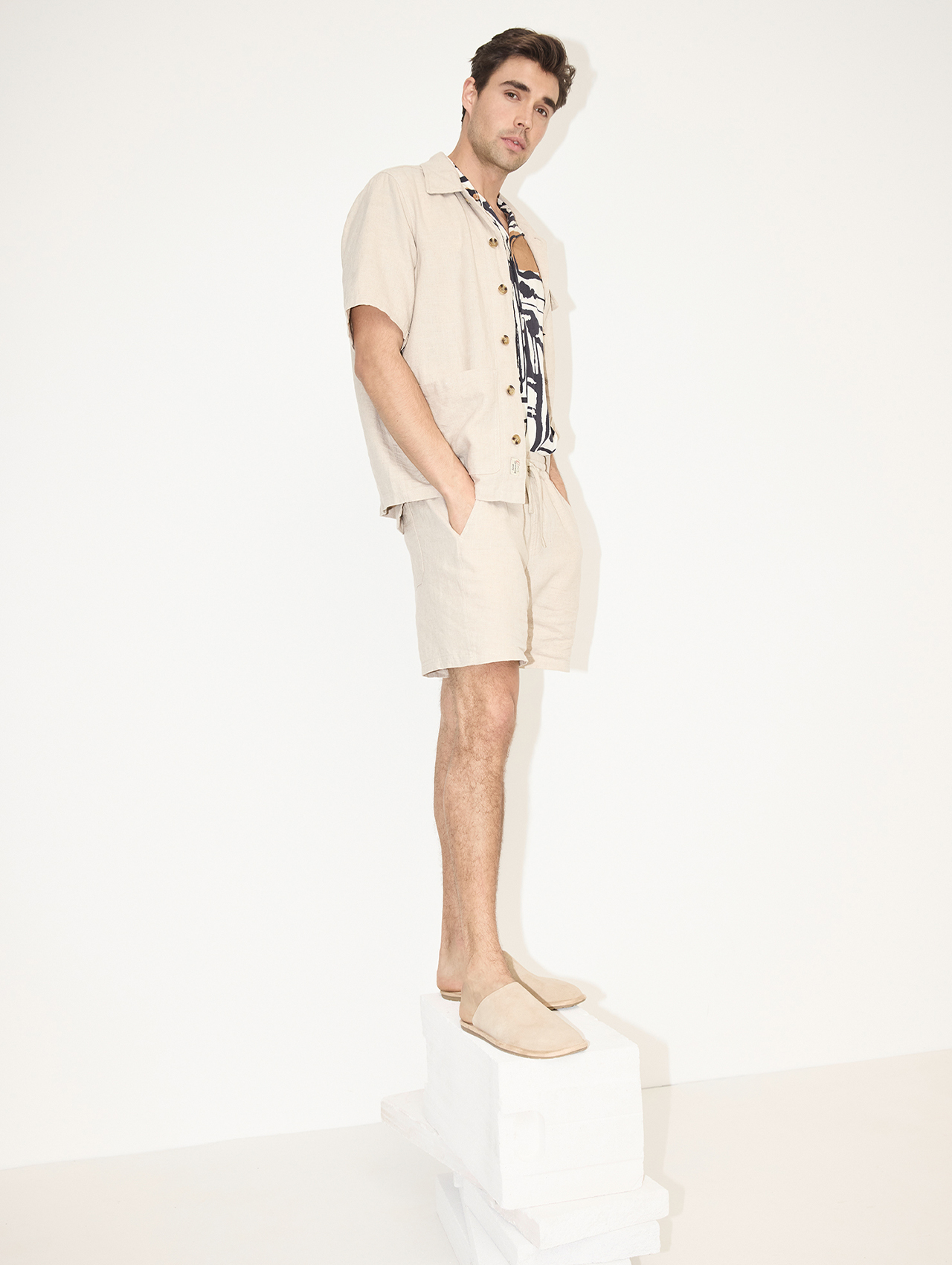 Ein Mann in einem beigen kurzärmeligen Hemd, passenden Shorts und Slippern steht auf einem weißen Podest vor einem schlichten weißen Hintergrund. Er blickt mit entspanntem Gesichtsausdruck leicht zur Seite.
