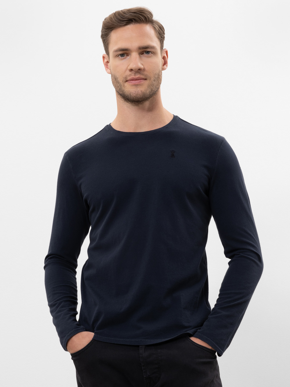 Ein Mann mit kurzen braunen Haaren, der ein tailliertes, langärmeliges, dunkles, marineblaues Hemd und eine schwarze Hose trägt, steht mit den Händen in den Taschen und blickt selbstbewusst in die Kamera vor einem schlichten weißen Hintergrund.