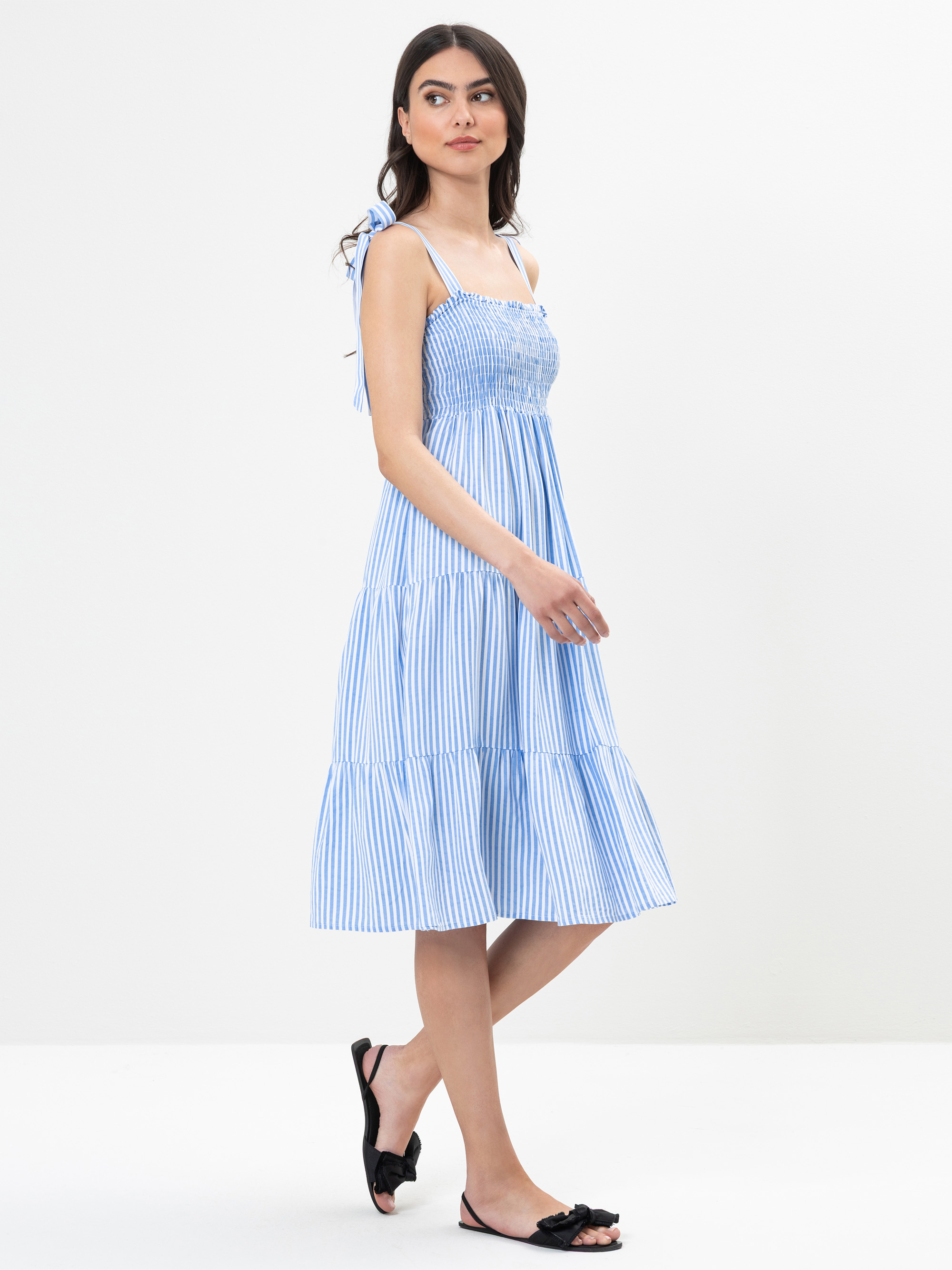 Frau in einem hellblau-weiß gestreiften, ärmellosen, knielangen Sommerkleid mit Bindebändern, schwarze flache Sandalen, stehend vor einem schlichten weißen Hintergrund.