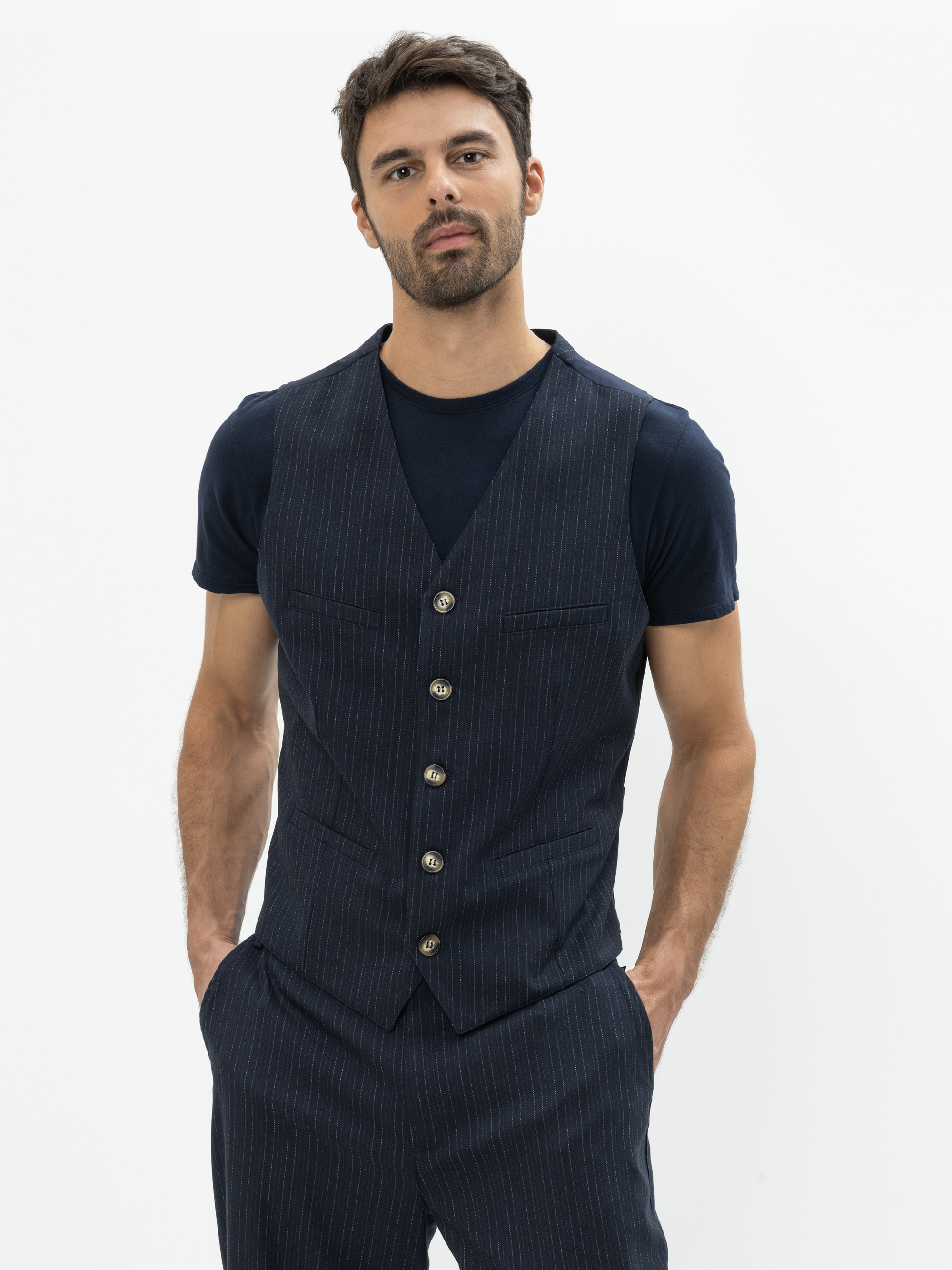 Ein Mann mit kurzen dunklen Haaren und einem Bart trägt eine marineblaue Nadelstreifenweste über einem marineblauen T-Shirt und einer passenden Nadelstreifenhose. Er steht mit den Händen in den Taschen vor einem schlichten weißen Hintergrund.