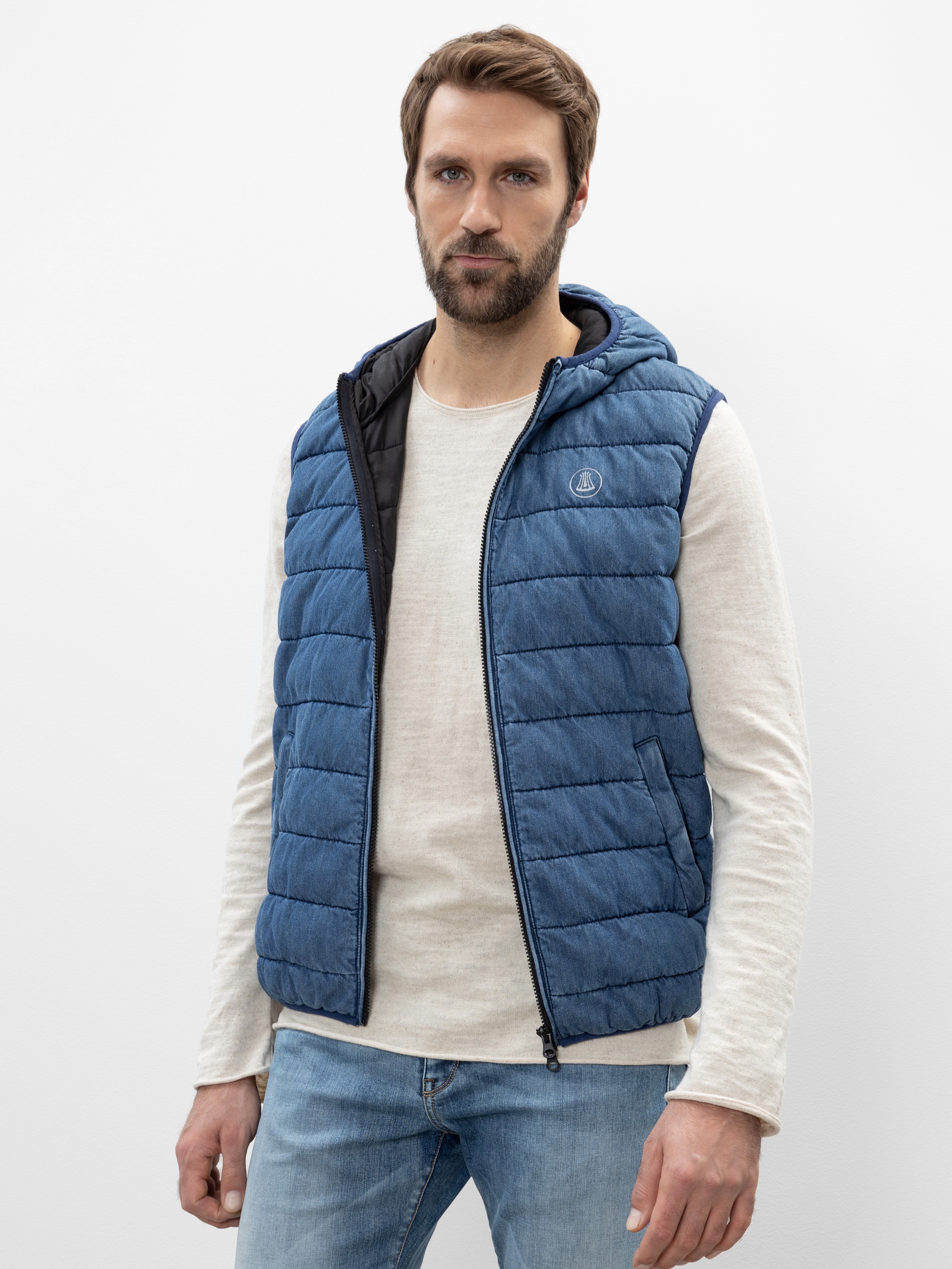 Ein Mann mit kurzen braunen Haaren und einem Bart trägt eine blaue Steppweste über einem hellbeigen, langärmeligen Hemd und blaue Jeans und steht vor einem schlichten weißen Hintergrund.