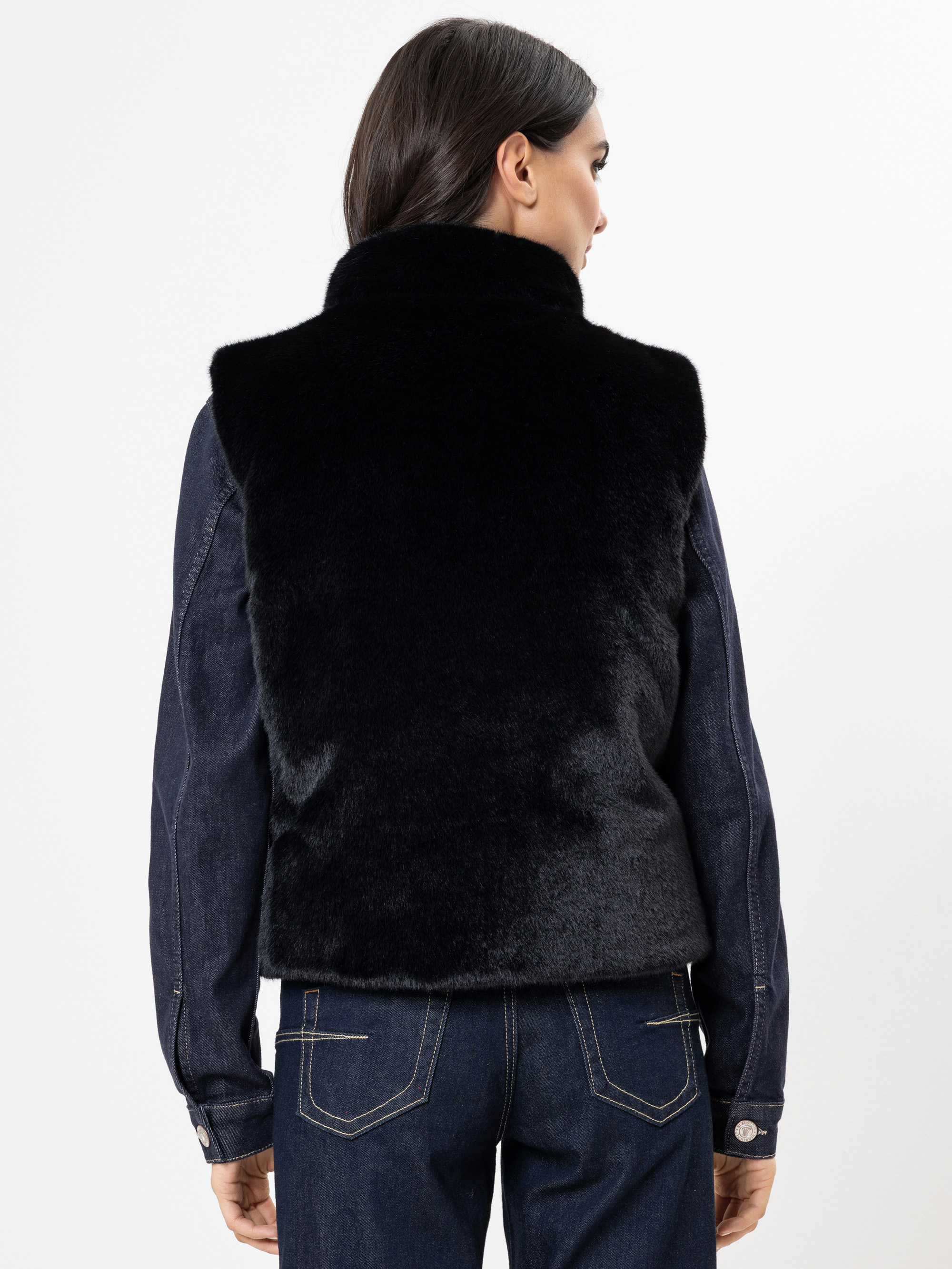 Eine Frau wird von hinten gezeigt, die eine schwarze Kunstpelzweste über einer dunklen Jeansjacke und dazu passende dunkle Jeans trägt. Sie steht vor einem schlichten weißen Hintergrund.