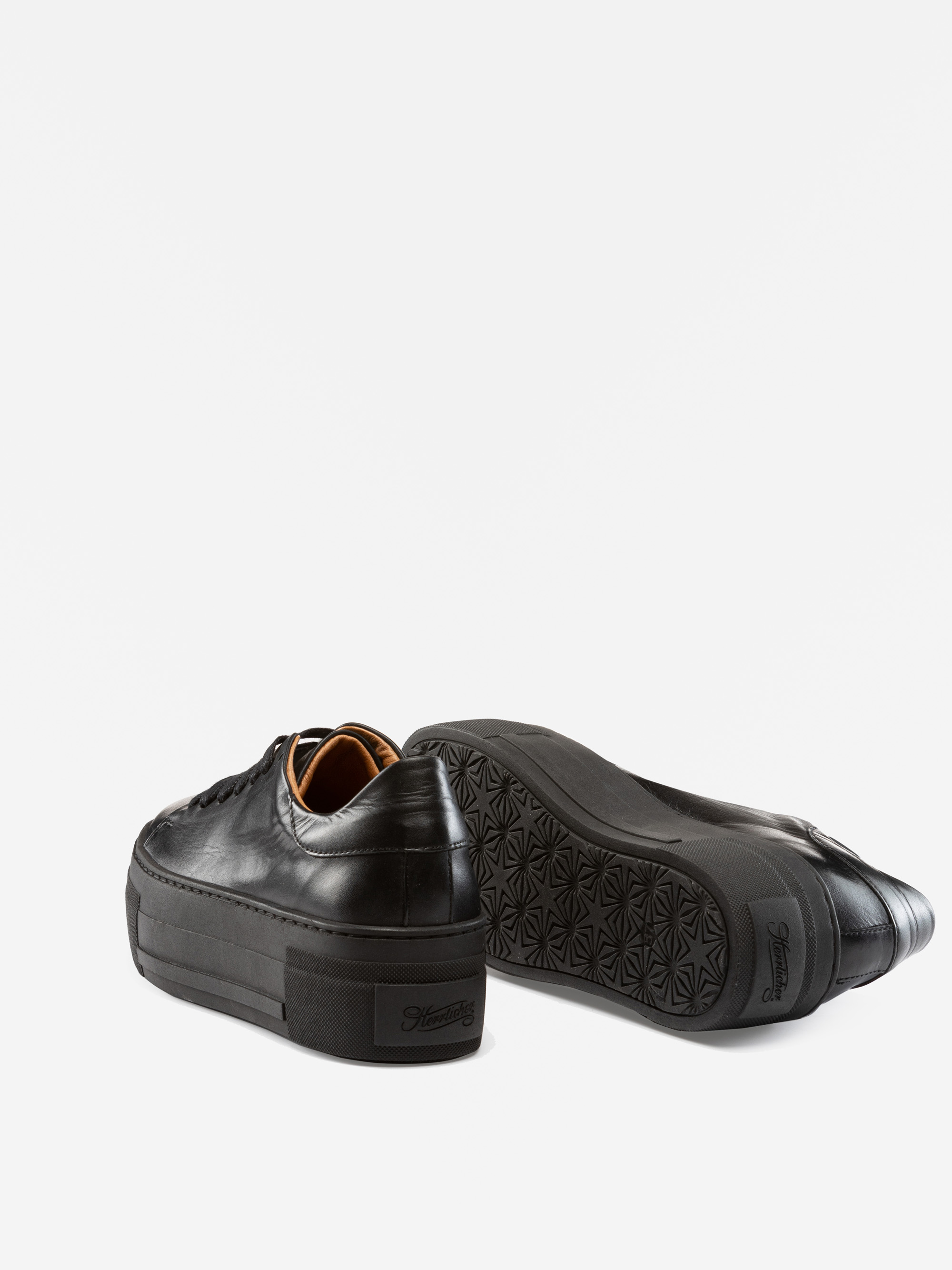 Ein Paar schwarzer Plateau-Sneakers aus Leder, von denen einer aufrecht steht und der andere auf der Seite liegt und die gemusterte Sohle zeigt, dargestellt auf weißem Hintergrund.