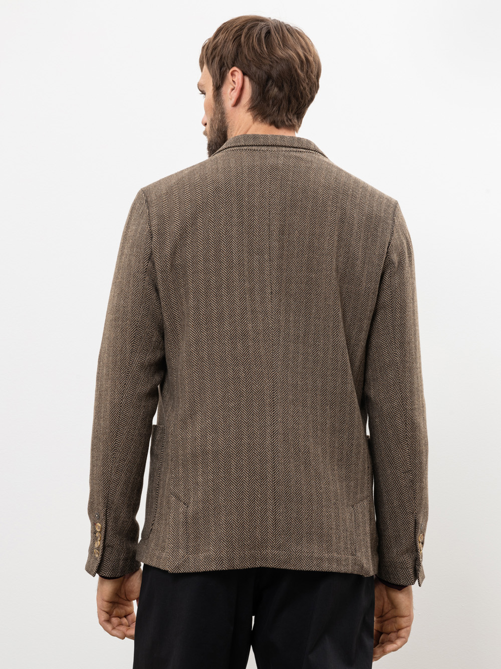 Ein Mann mit kurzen braunen Haaren wird von hinten gezeigt. Er trägt einen braunen Blazer mit Fischgrätenmuster und eine schwarze Hose und steht vor einem schlichten weißen Hintergrund.