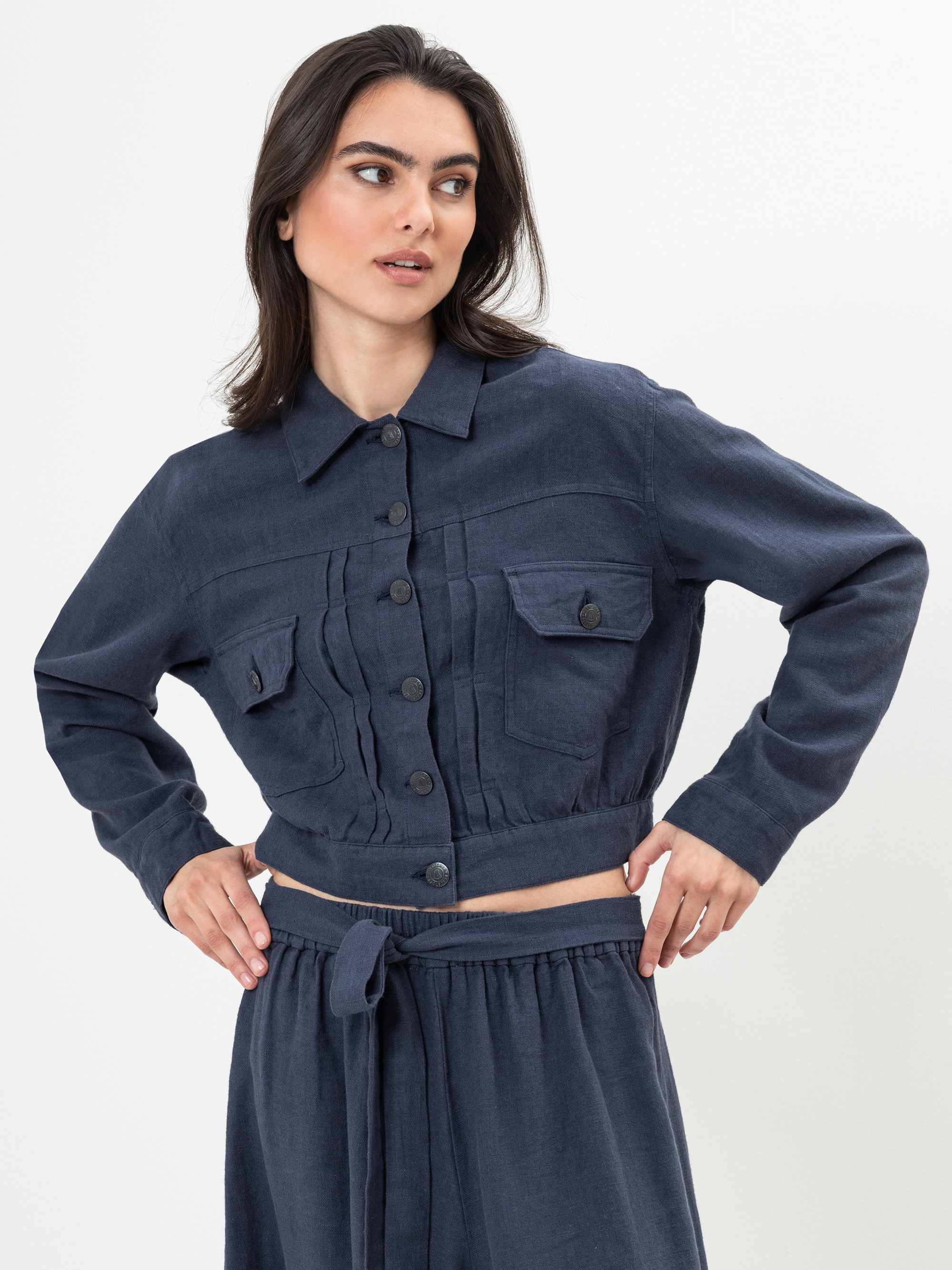 Eine Frau, die eine marineblaue, langärmelige, geknöpfte Jacke und eine dazu passende, hoch taillierte Hose mit Bindeband trägt, steht vor einem weißen Hintergrund und schaut mit einer Hand an der Hüfte leicht nach rechts.