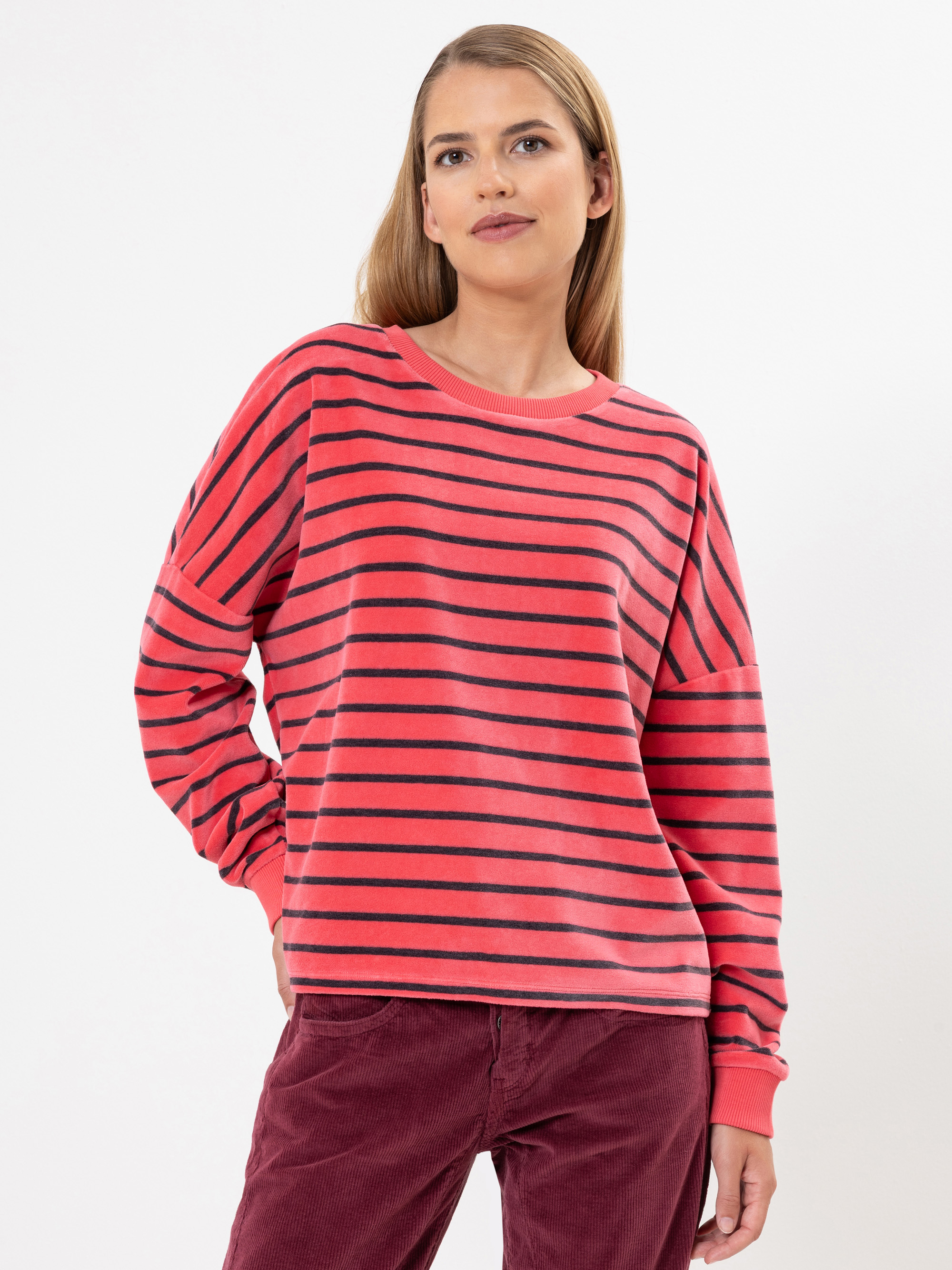 Eine Person mit langen, glatten blonden Haaren trägt einen rot-schwarz gestreiften Pullover und eine kastanienbraune Hose und steht vor einem schlichten weißen Hintergrund.