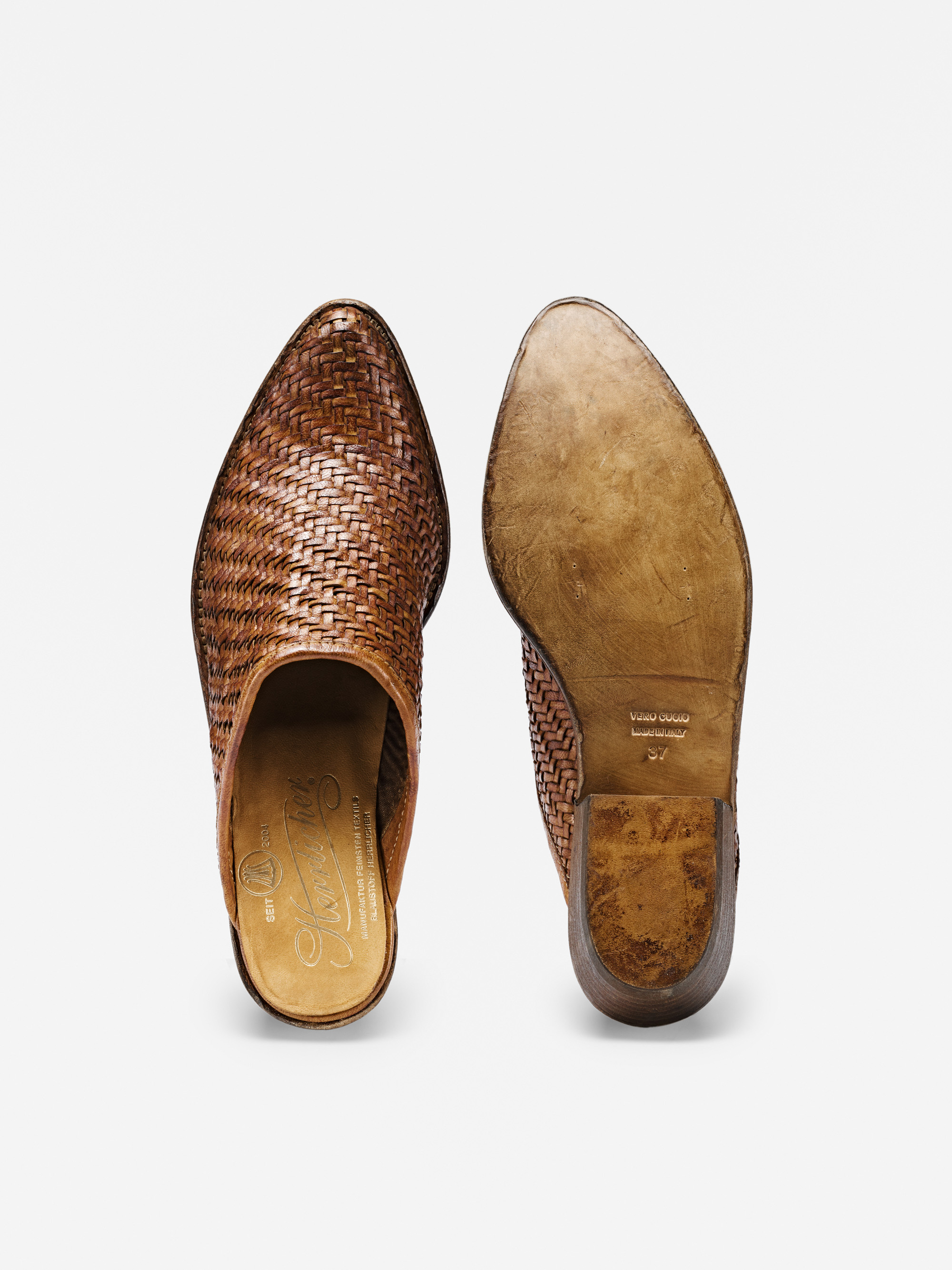 Ein Paar brauner Slipper aus gewebtem Leder, dargestellt von oben und von der Sohle aus, vor einem weißen Hintergrund. Der linke Schuh zeigt das gewebte Detail, während der rechte Schuh die abgenutzte Ledersohle zeigt.