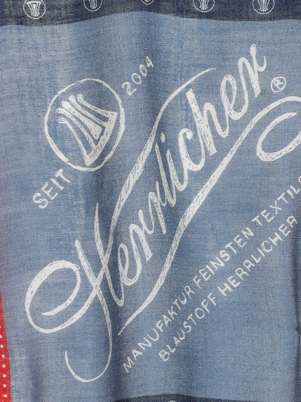 Auf einem Stoffetikett mit weißer Schrift steht "Seit 2004 Herrlicher Manufaktur Feinster Textil Blaustoff Herrlicher" auf blauem Hintergrund, mit einem kreisförmigen Logo und Ziernähten.