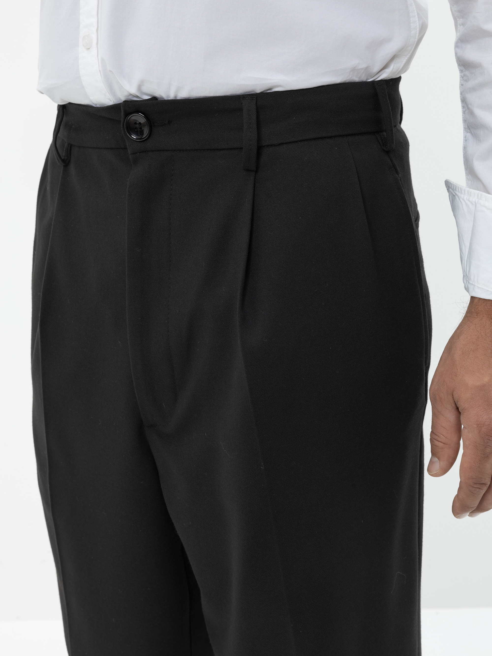 Eine Person, die ein weißes Hemd trägt, das in eine schwarze formelle Hose mit geknöpftem Bund gesteckt ist, wird von der Taille abwärts vor einem schlichten Hintergrund gezeigt.