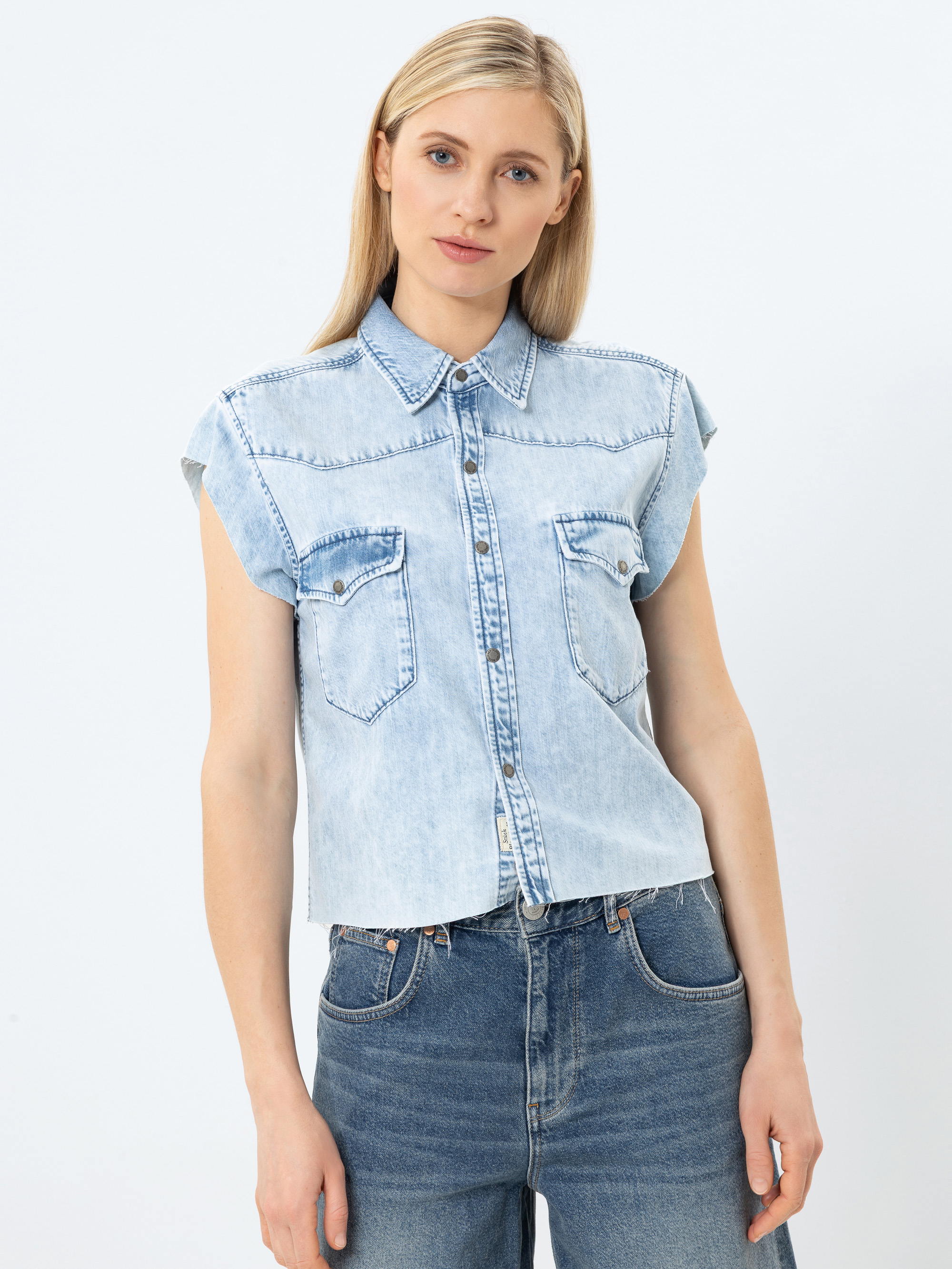 Eine Frau mit langen blonden Haaren trägt ein ärmelloses, hellblaues Jeanshemd mit Vordertaschen und eine blaue Jeans, die vor einem schlichten weißen Hintergrund steht.
