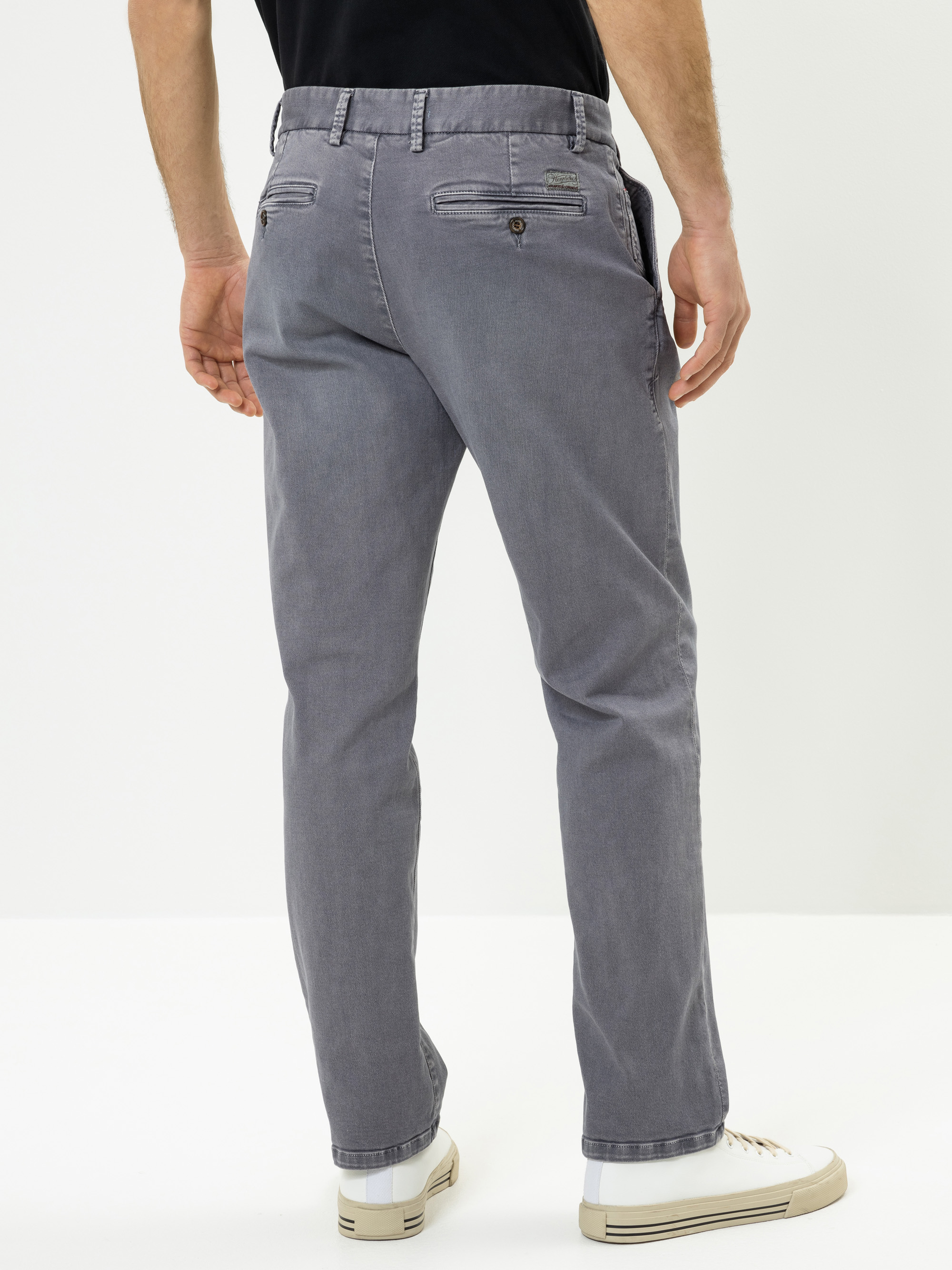 Ein Mann in grauen Chinos und weißen Turnschuhen steht mit dem Rücken zur Kamera vor einem schlichten weißen Hintergrund.