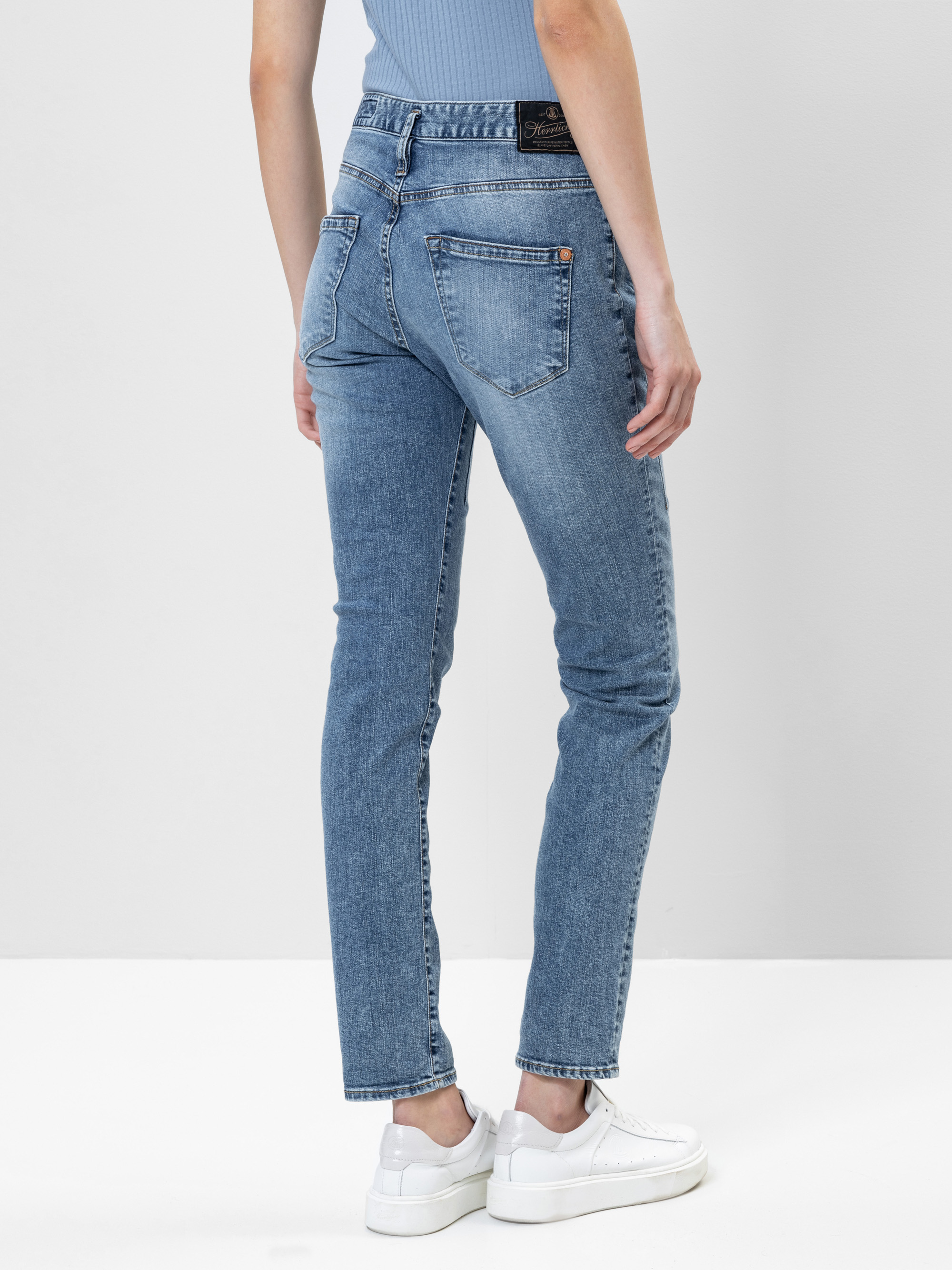 Eine Person, die eine hellblaue Jeans und weiße Turnschuhe trägt, steht mit dem Gesicht nach außen, den rechten Arm entspannt an der Seite, vor einem schlichten hellen Hintergrund.