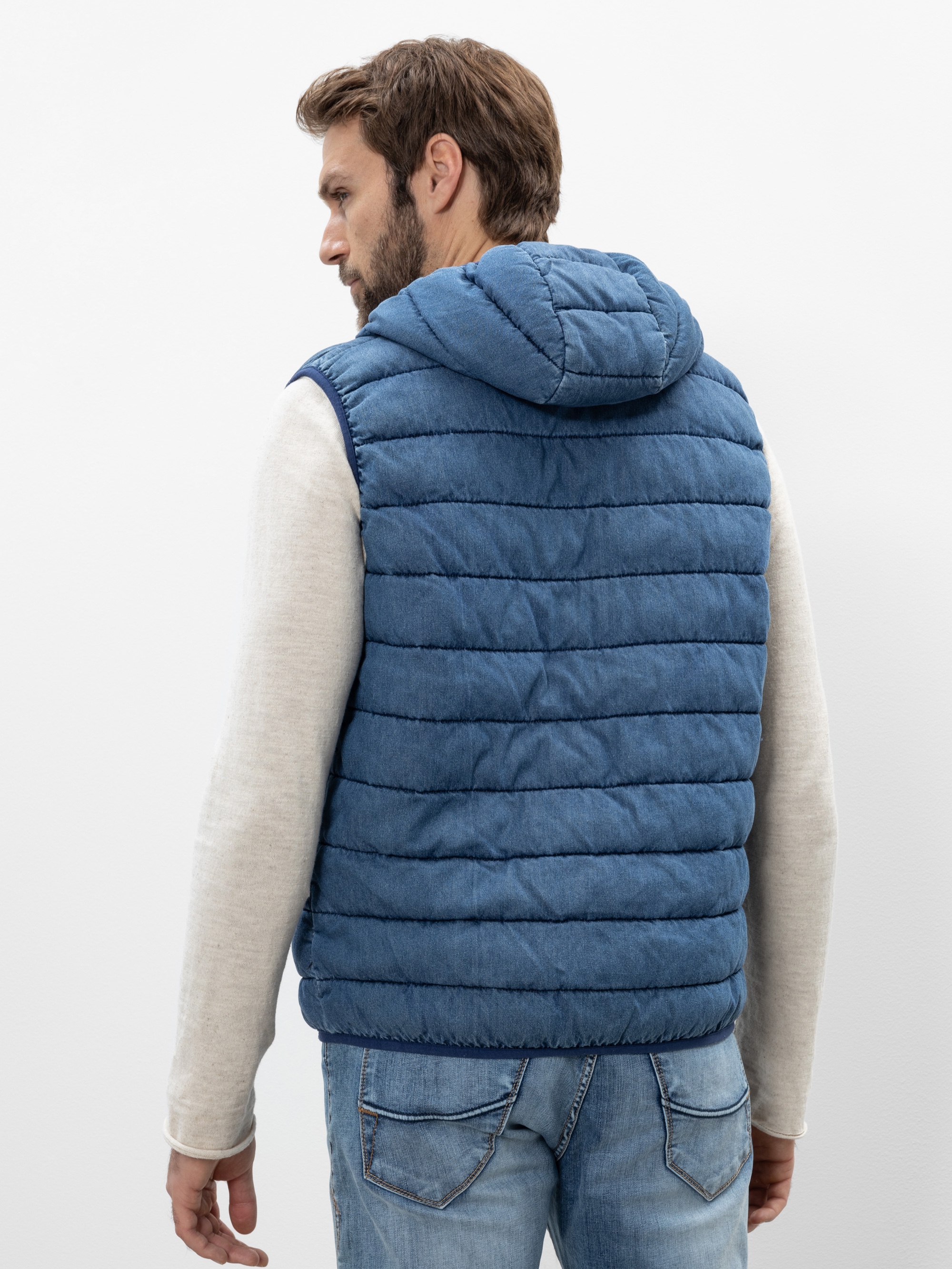 Von hinten ist ein Mann zu sehen, der eine blaue Steppweste mit Kapuze über einem hellen langärmeligen Hemd und blaue Jeans trägt und vor einem schlichten weißen Hintergrund steht.