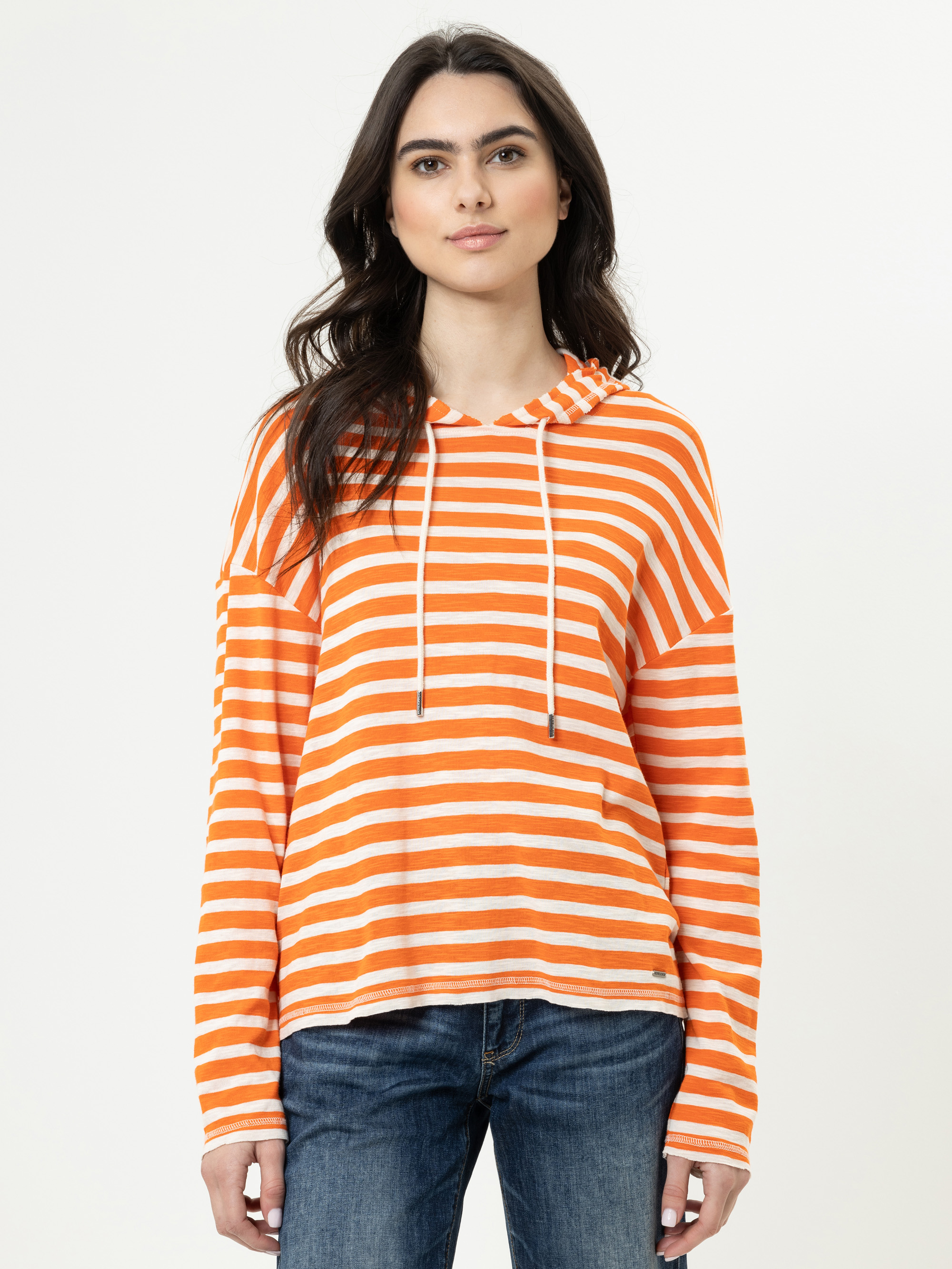 Eine Frau mit langen braunen Haaren trägt einen lässigen orange-weiß gestreiften Kapuzenpullover und blaue Jeans und steht vor einem schlichten weißen Hintergrund. Sie blickt nach vorne und hat einen neutralen Gesichtsausdruck.