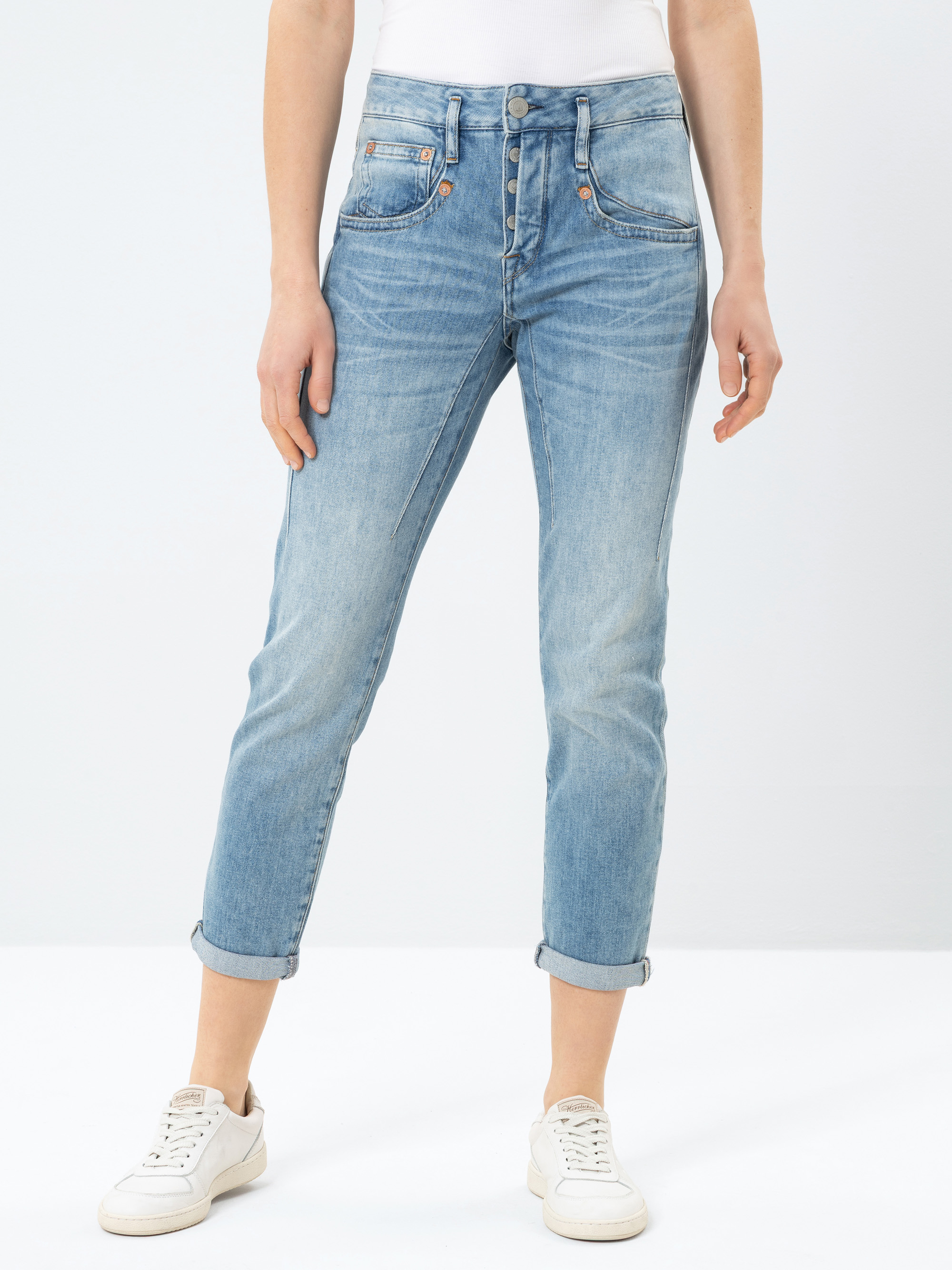 Eine Person, die hellblaue, hochgekrempelte Jeans, ein weißes Oberteil und weiße Turnschuhe trägt, steht vor einem schlichten weißen Hintergrund.