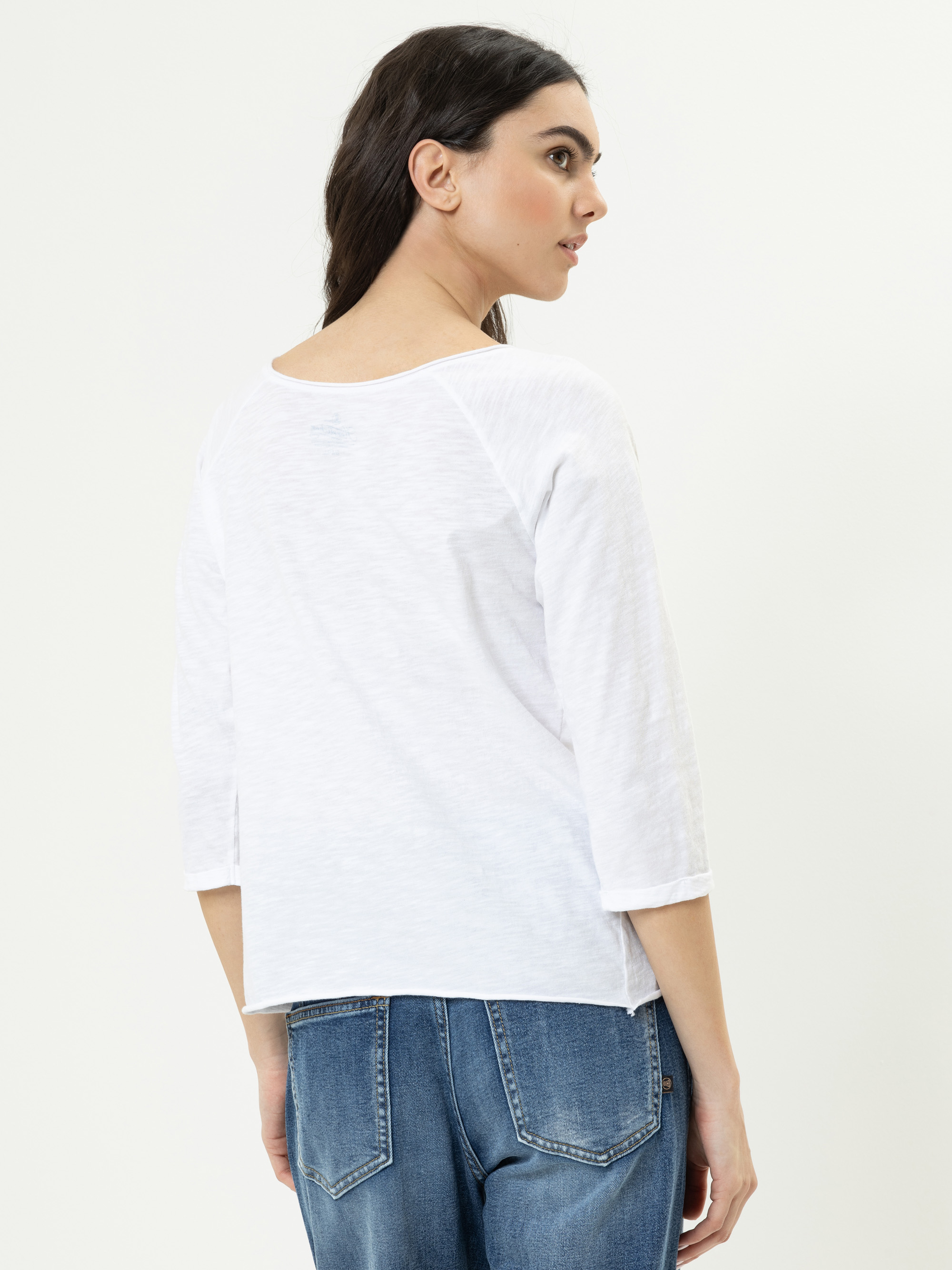 Eine Frau mit langen dunklen Haaren trägt ein weißes dreiviertelärmeliges Oberteil und blaue Jeans. Sie steht und schaut über ihre linke Schulter vor einem schlichten hellen Hintergrund.