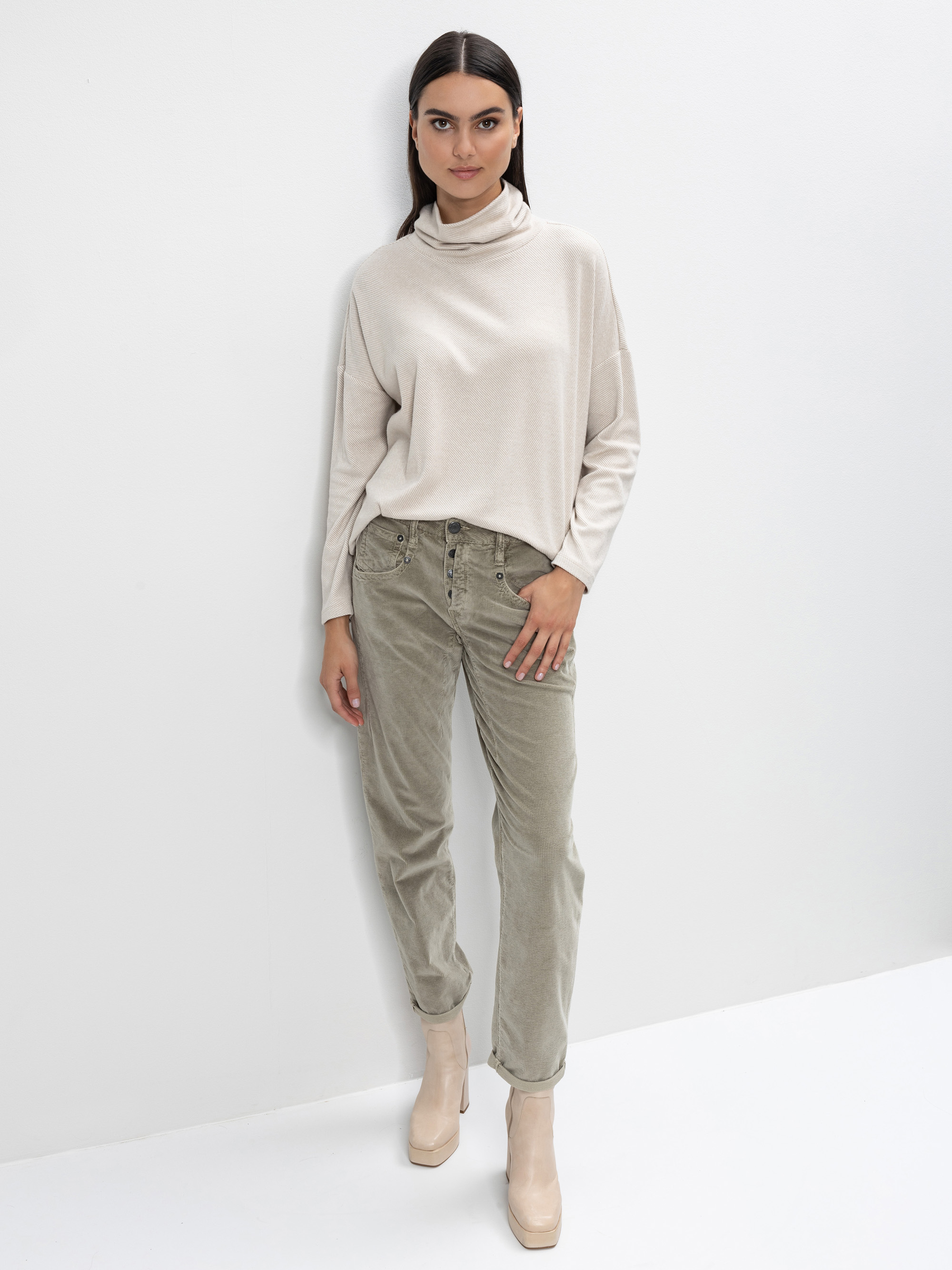 Eine Frau mit langen dunklen Haaren steht vor einer weißen Wand. Sie trägt einen hellbeigen Rollkragenpullover, eine hellolivgrüne Kordhose und beigefarbene Stiefel mit Absätzen. Sie blickt mit neutralem Blick in die Kamera.