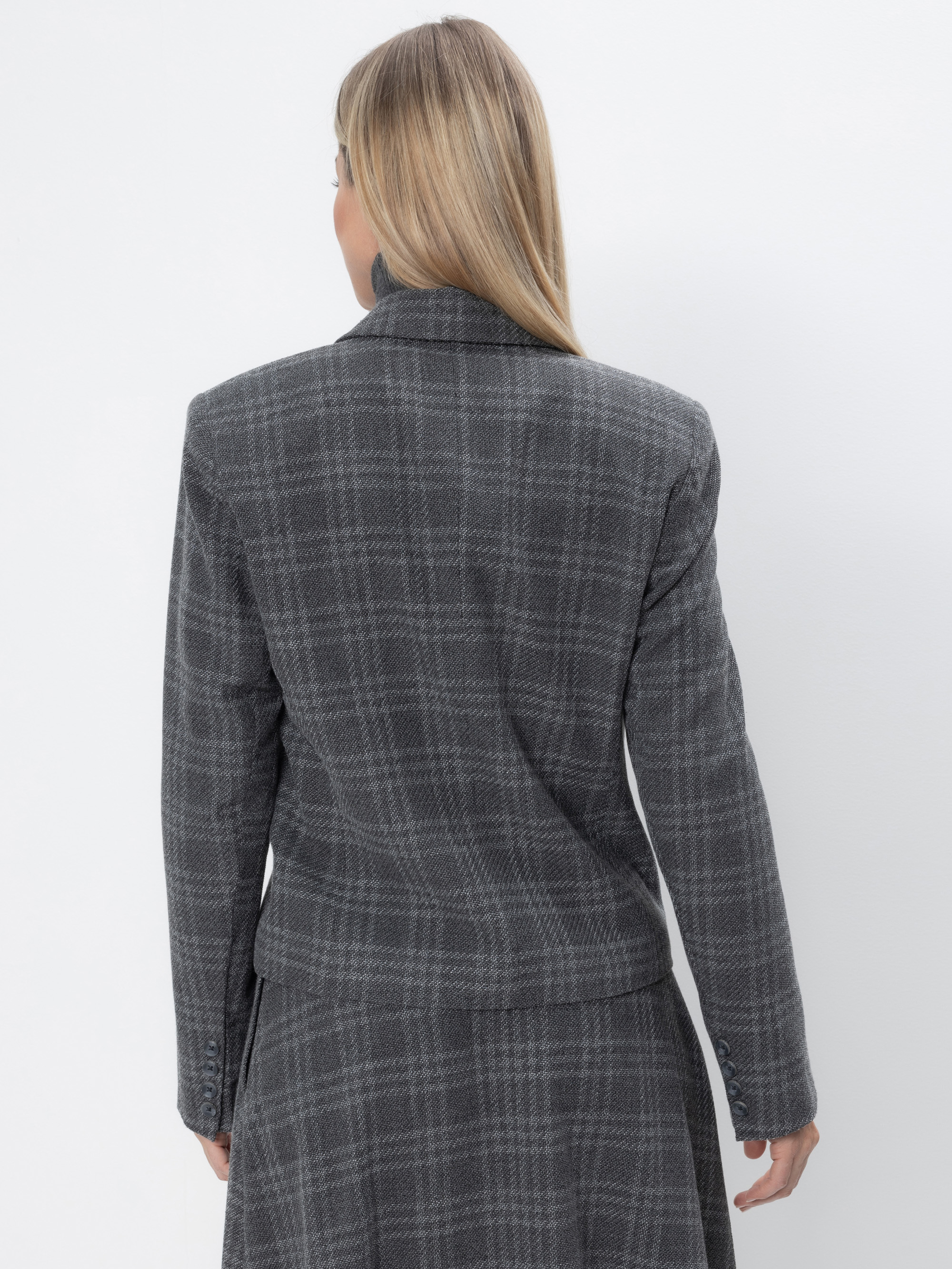 Eine Person mit langen blonden Haaren, von hinten gesehen, trägt einen grau karierten Blazer mit passendem Rock. Der Hintergrund ist einfarbig weiß.