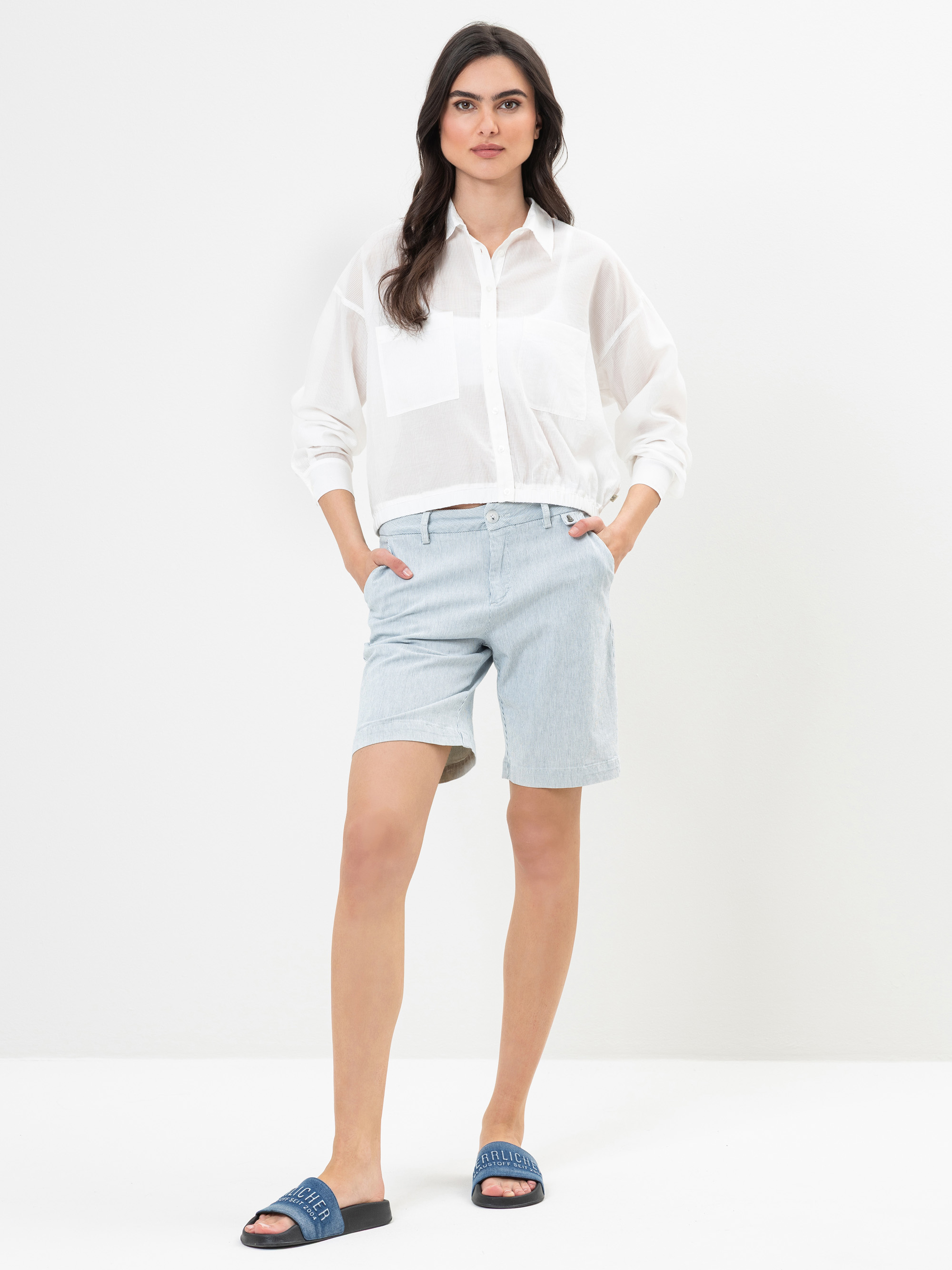 Eine Frau steht vor einem weißen Hintergrund und trägt ein durchsichtiges weißes langärmeliges Hemd, hellblaue knielange Shorts und blaue Slipper-Sandalen. Ihre Hände stecken in den Taschen und sie hat langes dunkles Haar.