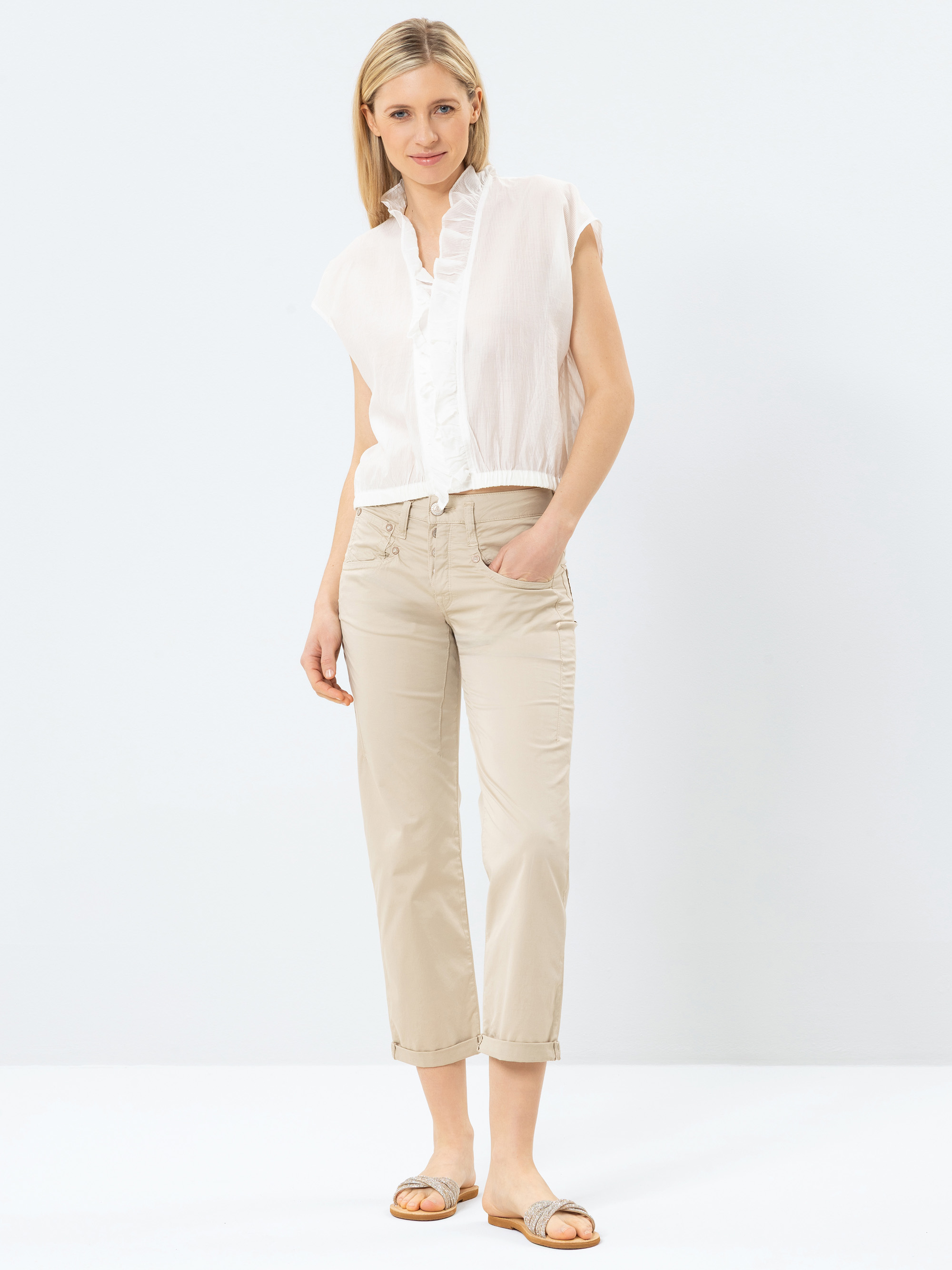 Eine Frau steht vor einem schlichten Hintergrund und trägt eine weiße, kurzärmelige Bluse mit Rüschenkragen, eine beigefarbene, kurze Hose und hellbraune Sandalen. Sie hat glattes blondes Haar und lächelt sanft mit einer Hand in ihrer Tasche.