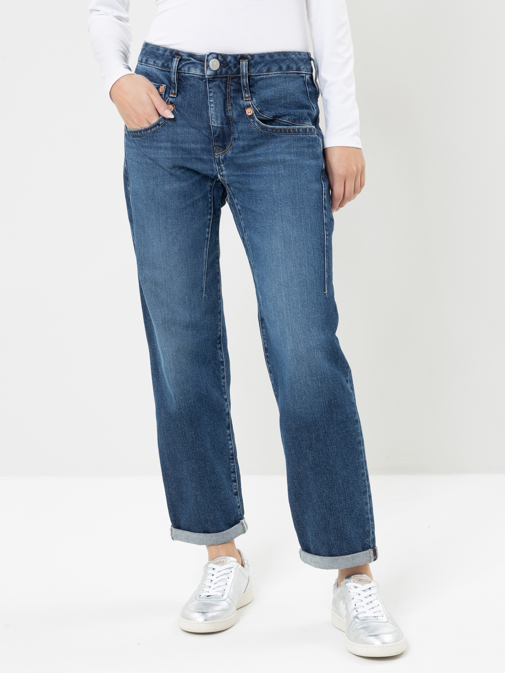 Eine Person, die eine blaue Jeans mit geradem Bein, ein weißes Langarm-Oberteil und silberne Sneaker trägt, steht vor einem schlichten, hellen Hintergrund und hat die Hände in den Taschen. Die Jeans ist an den Knöcheln leicht umgeschlagen. Der Kopf de